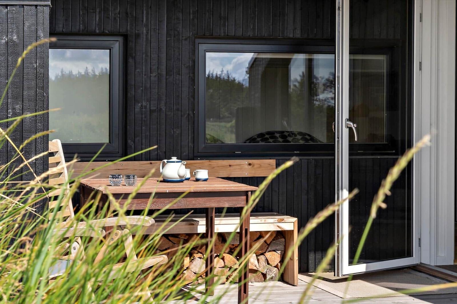 Terrasse mit Holztisch, Stühlen und Kaminholz vor einem Haus mit schwarzer Fassade.