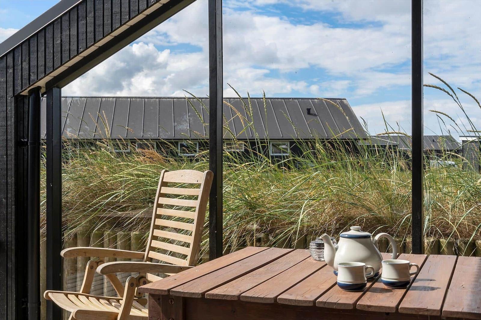 Holzstuhl und Tisch mit Teekanne und Tassen auf einer Terrasse mit Blick auf Gräser und ein Haus.