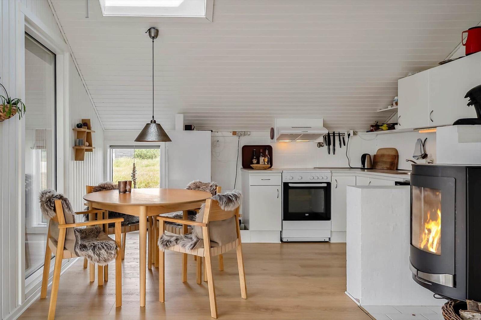 Küche mit Esstisch, Holzfußboden und Kaminofen. Weiße Schränke und Fenster mit Blick nach draußen.