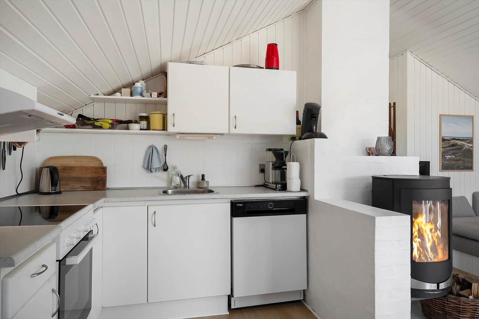 Küche mit weißen Schränken, Spüle und Waschmaschine. Kamin mit brennendem Holz.