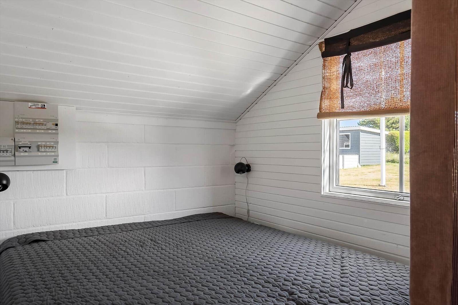 Schlafzimmer mit Dachboden, Bett, Fenster und Stromverteiler an der Wand.