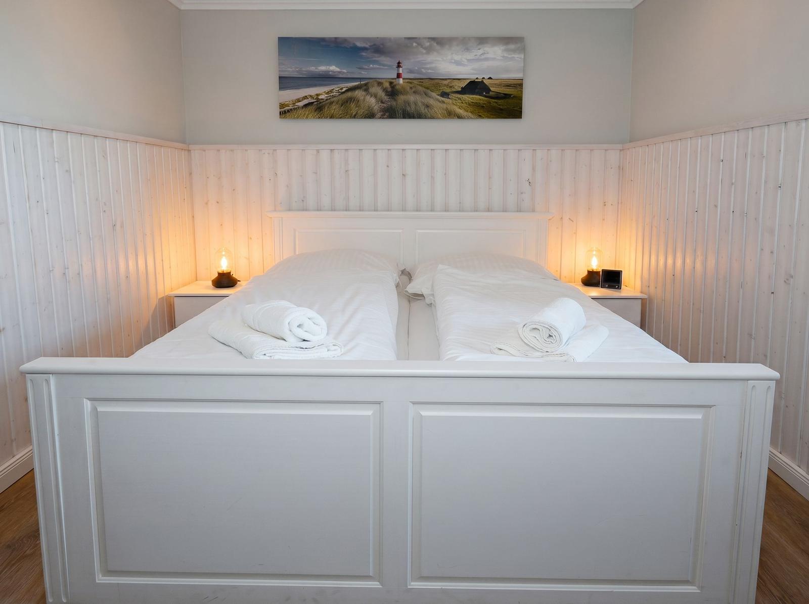 Schlafzimmer mit weißem Bett, Holzwandverkleidung und Bild an der Wand.