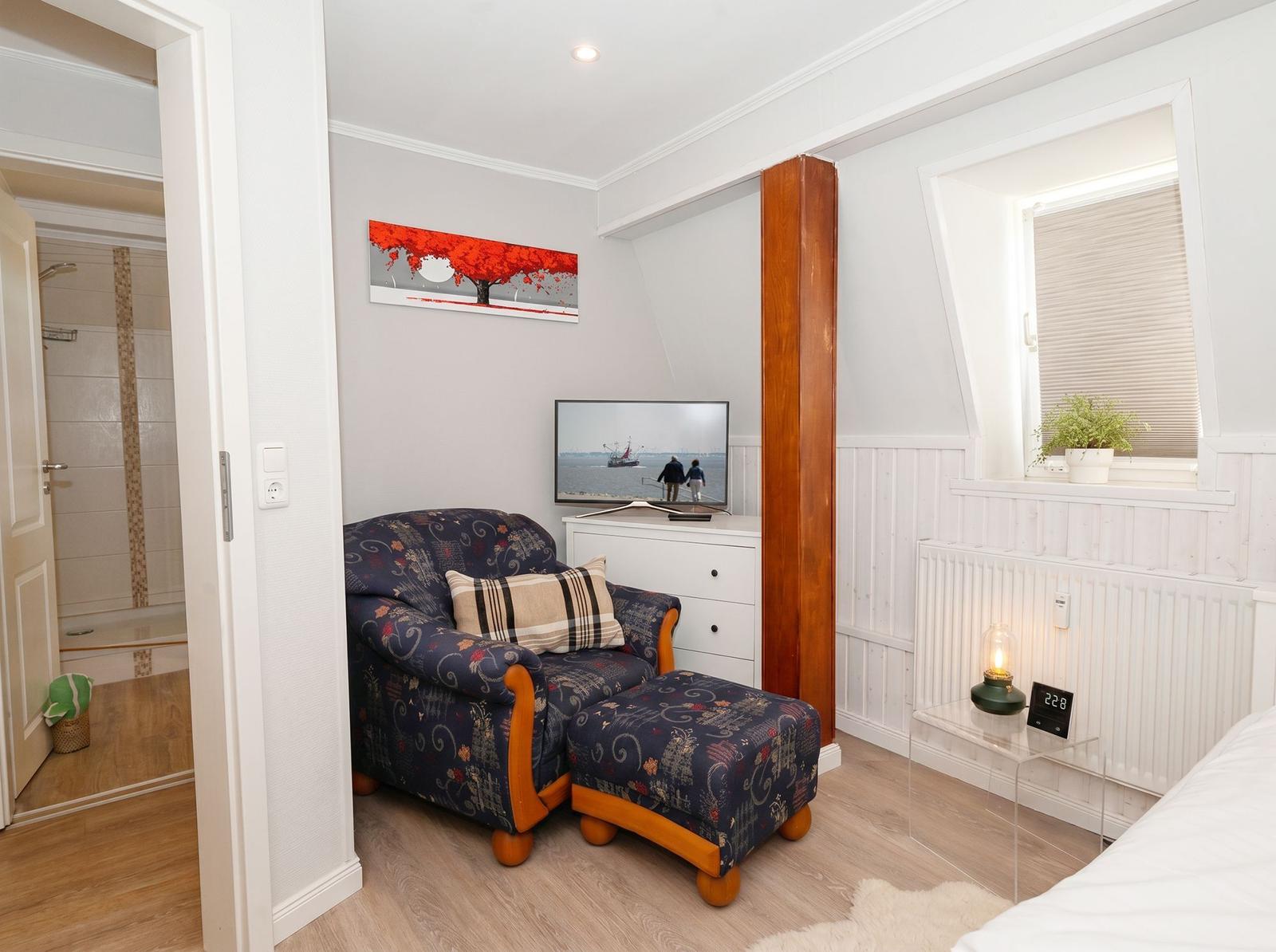Ein Raum mit Sessel, Fernseher und Blick in die Badewanne.