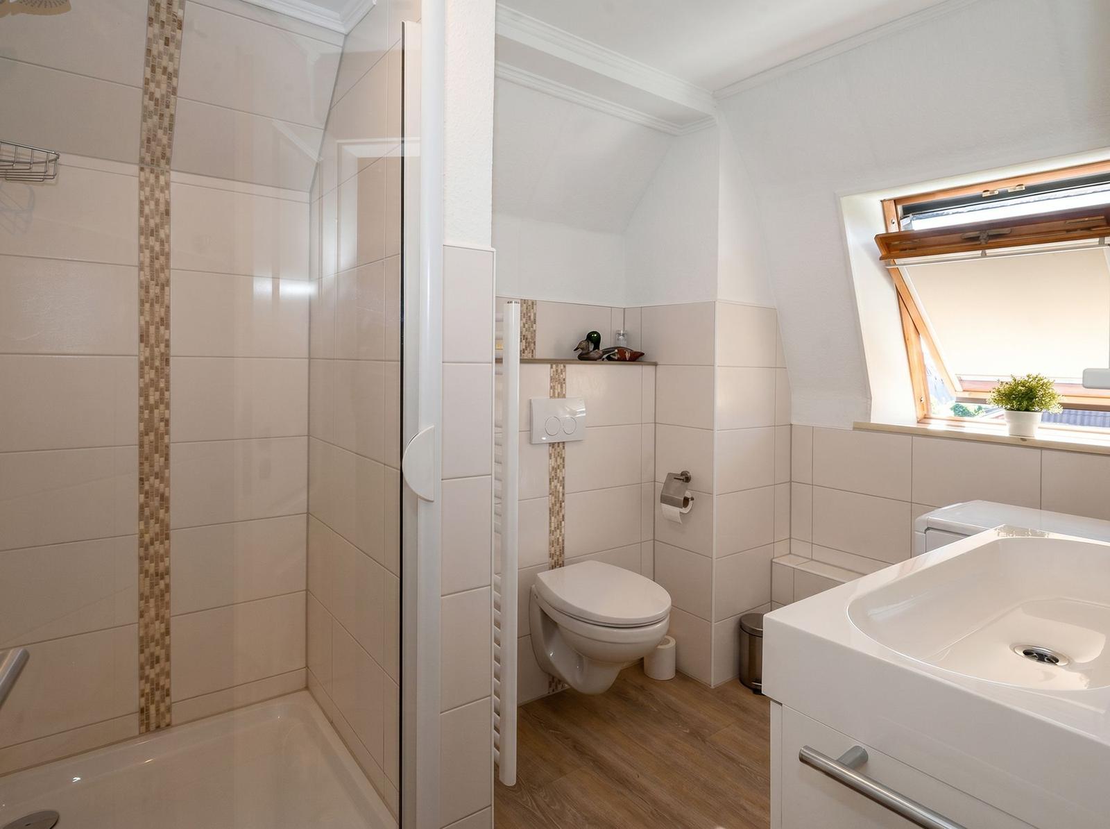 Badezimmer mit Dusche, Toilette und Waschbecken. Fenster mit Pflanze.