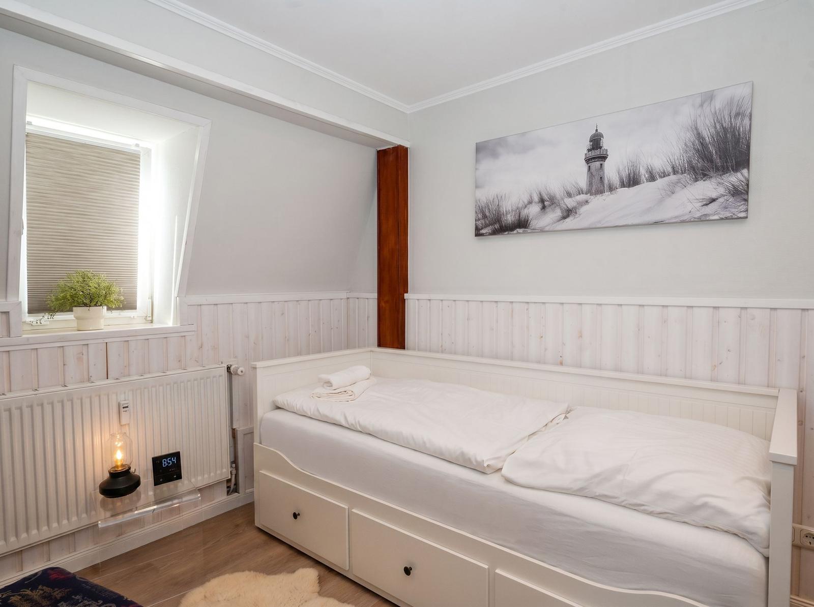 Schlafzimmer mit weißem Bett, Wandbild Leuchtturm und Fenster mit Jalousie.