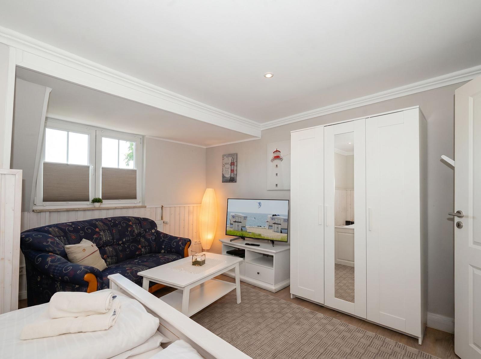 Wohnzimmer mit Sofa, Fernseher und Kleiderschrank. Fenster mit Jalousien und Teppichboden.