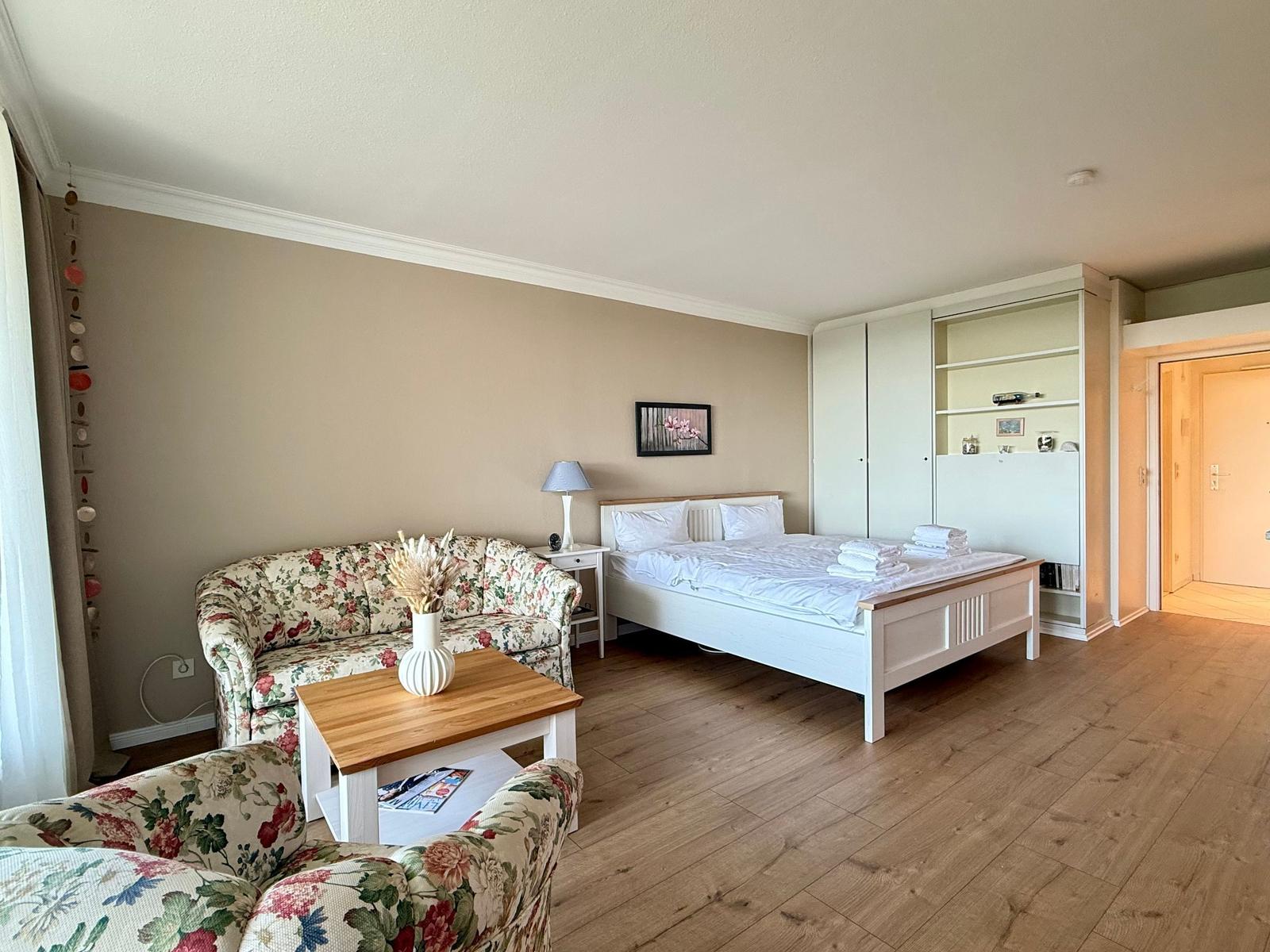 Room with bed, sofa, table, and closet. Wooden floor and beige walls.
