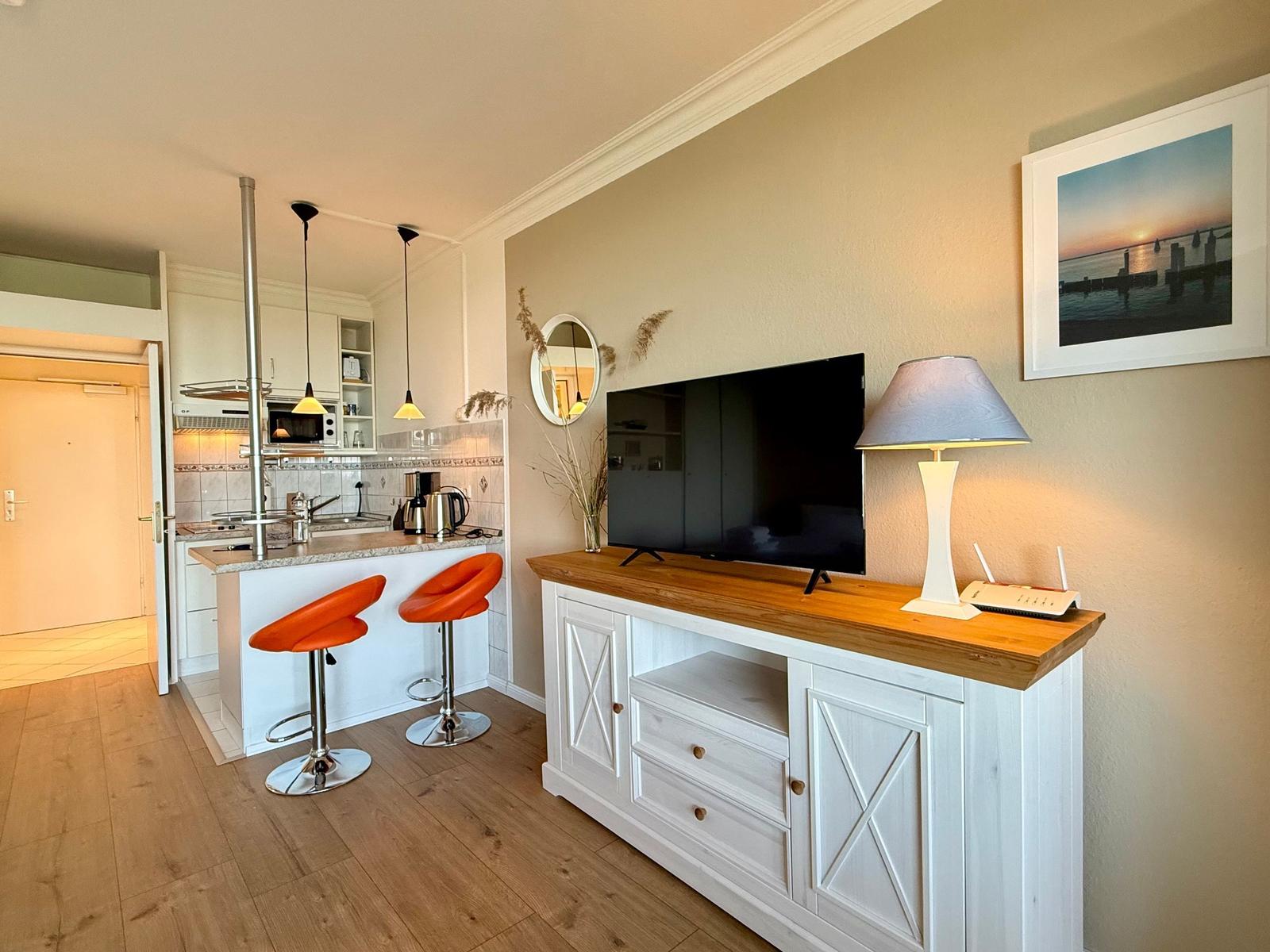 Living area with TV stand, lamp, and kitchen area with orange bar stools.
