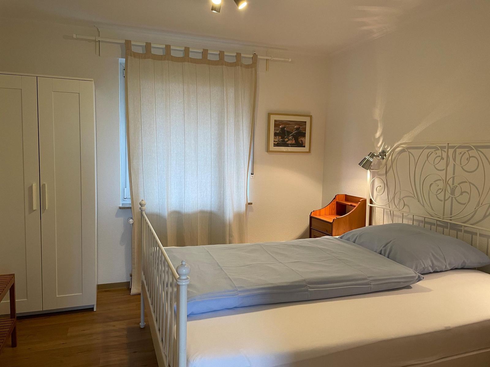 Schlafzimmer mit weißem Metallbett, Holzschrank und Vorhang vor Fenster.