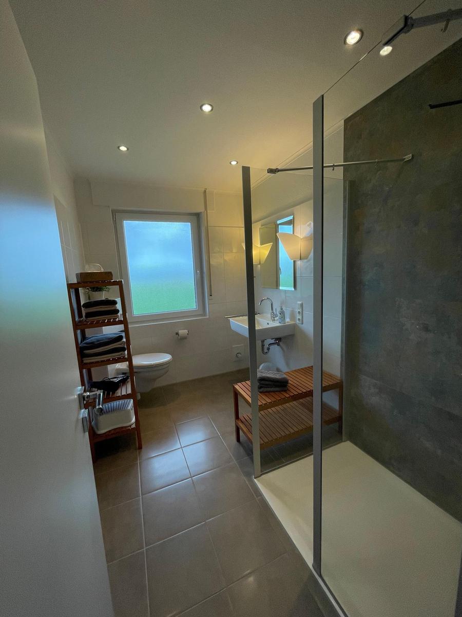 Badezimmer mit Dusche, Waschbecken und Toilette. Fenster mit Blick nach draußen.