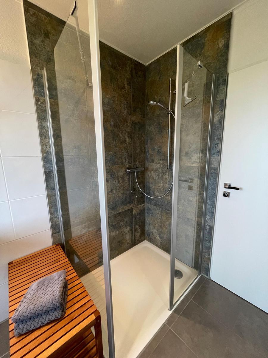 Dusche mit Glaswand, dunklen Fliesen und Holzbank mit Handtuch.