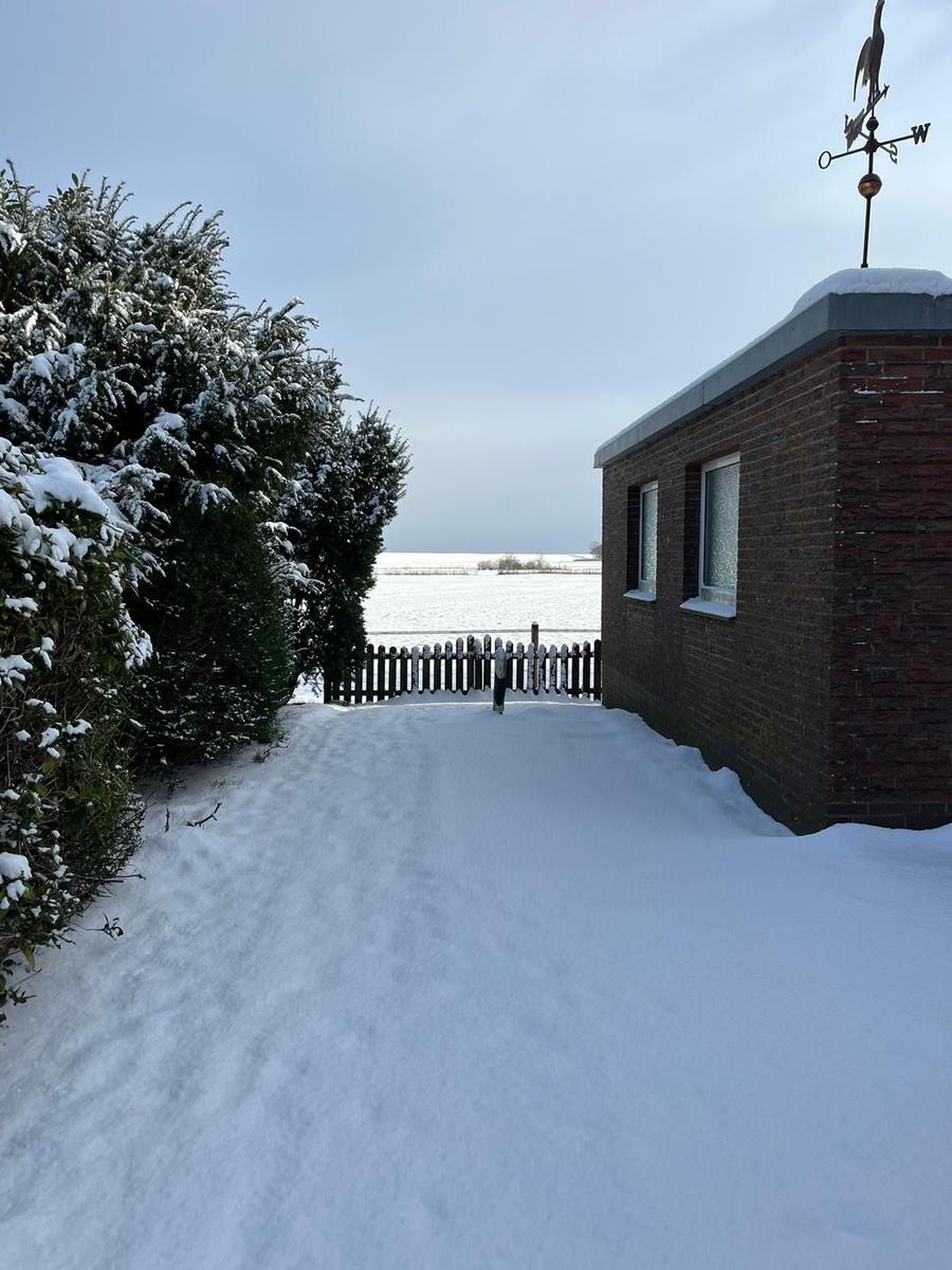 Schneebedeckter Weg führt zu einer kleinen Hütte mit Windschutz und Blick auf eine winterliche Landschaft.