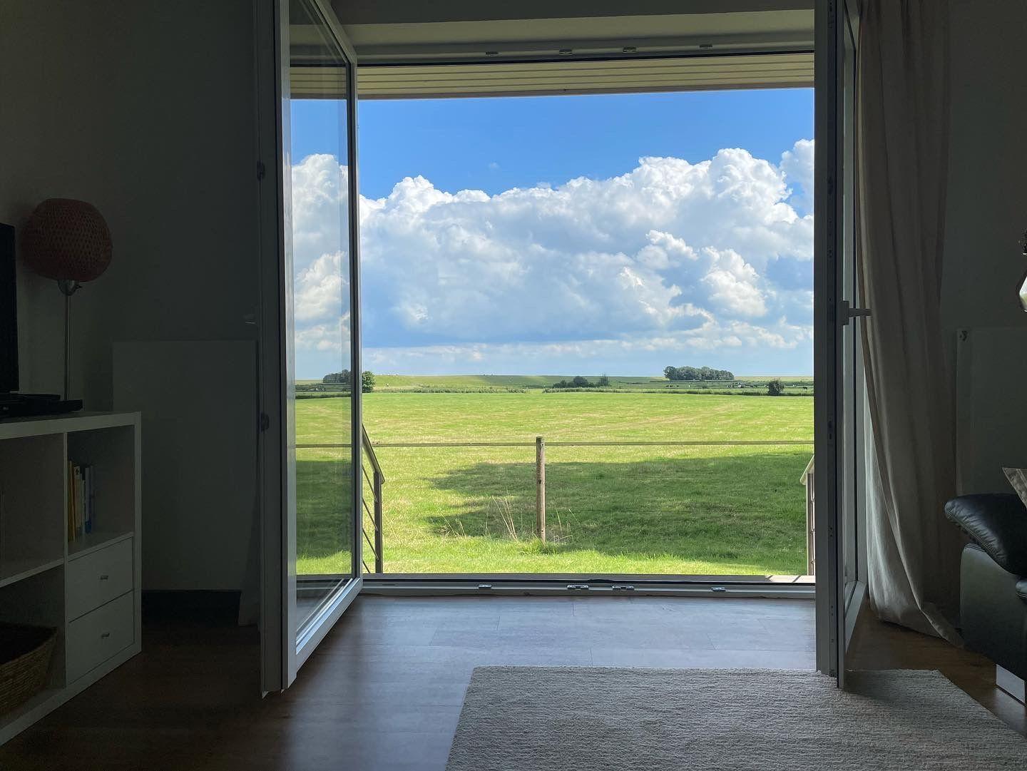 Durch die Glastüren blickt man auf eine grüne Wiese unter blauem Himmel mit weißen Wolken.