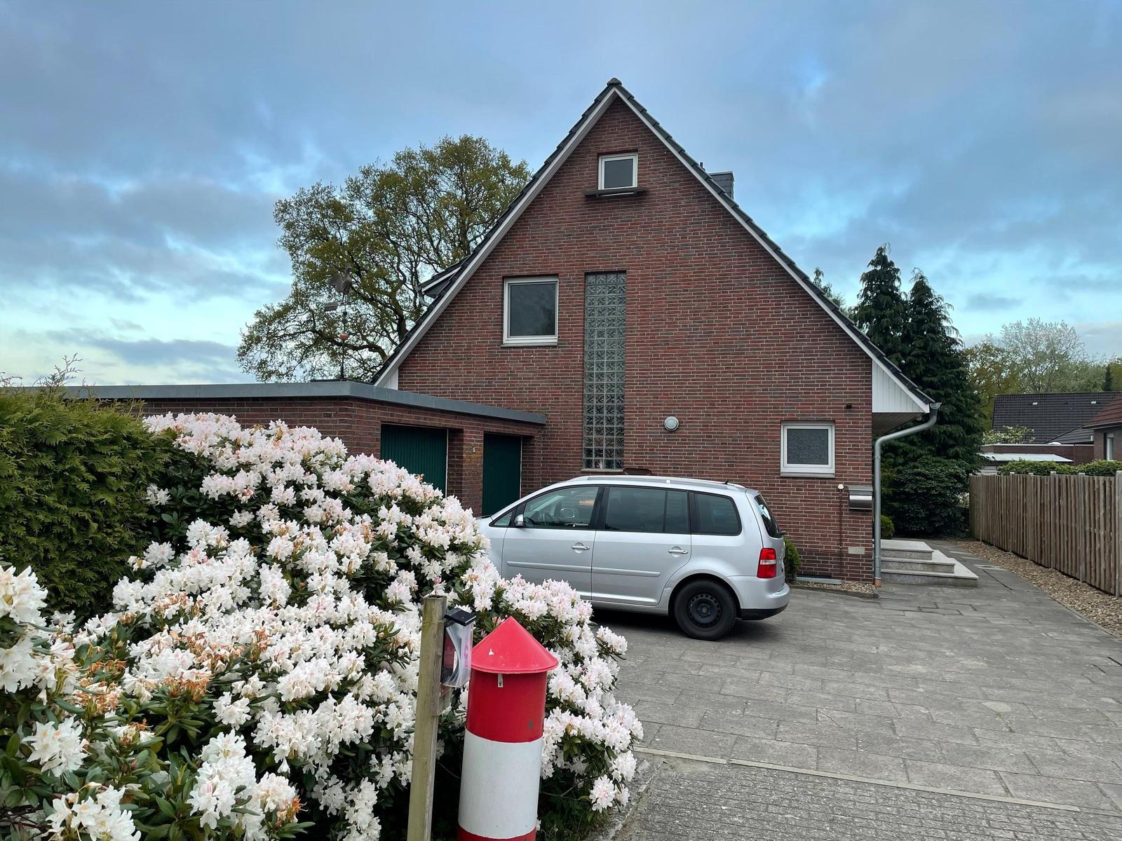 Rotes Backsteinhaus mit Parkplatz und blühenden Sträuchern im Vorgarten.