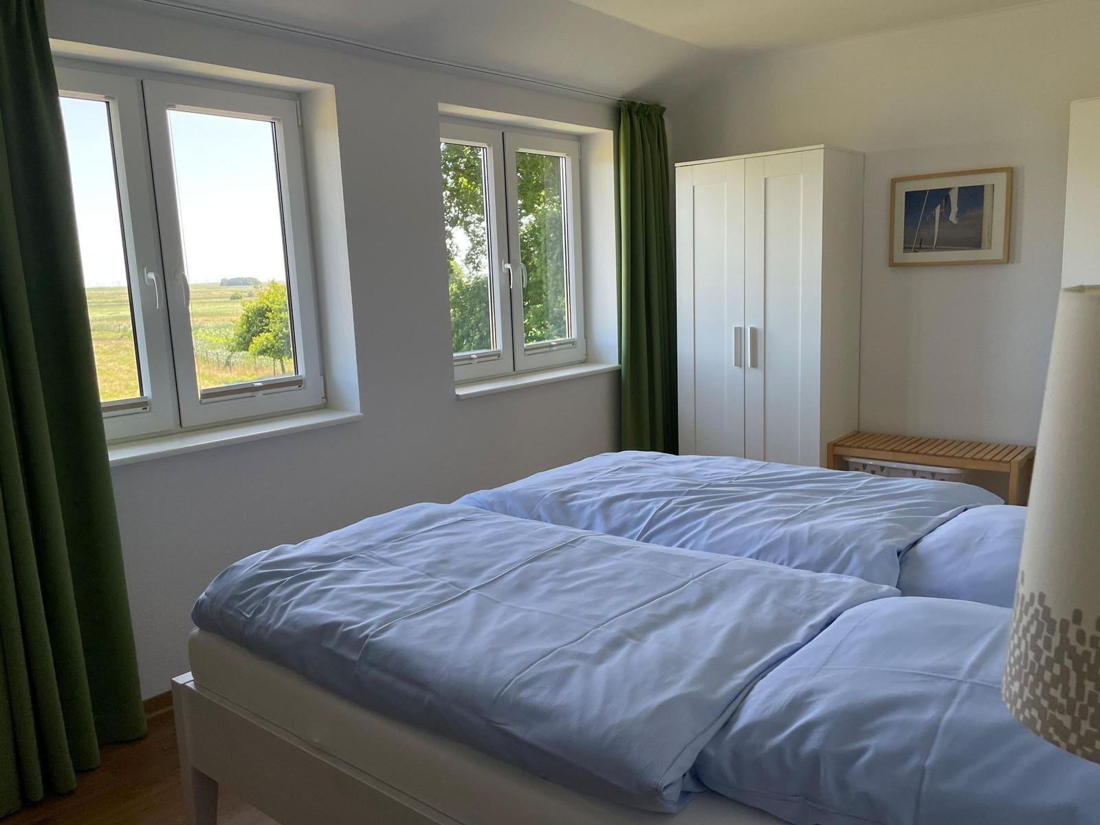 Schlafzimmer mit Doppelbett, zwei Fenstern und weißem Schrank.