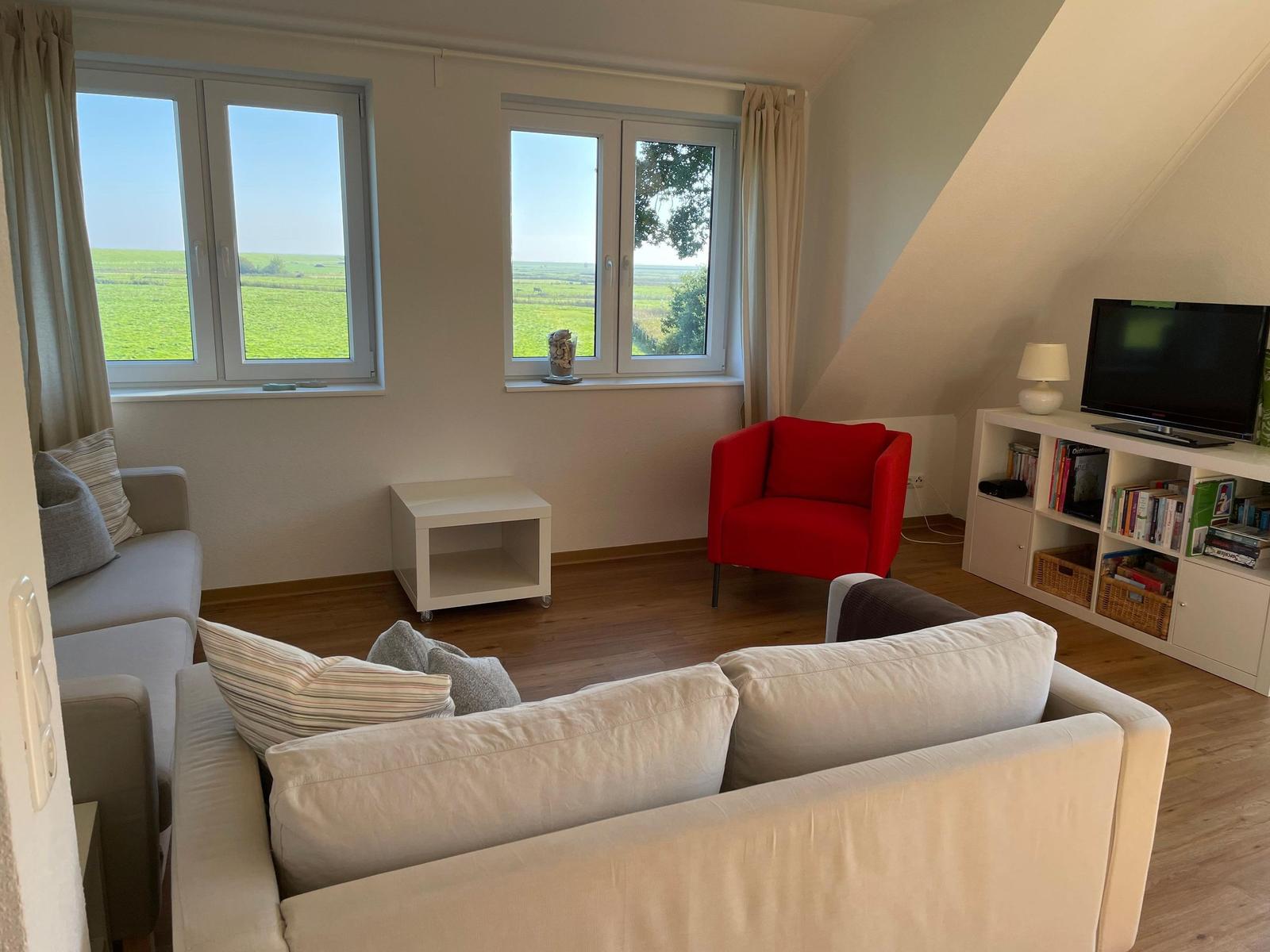 Wohnzimmer mit Sofa, rotem Sessel und TV. Fenster zeigen grüne Landschaft.