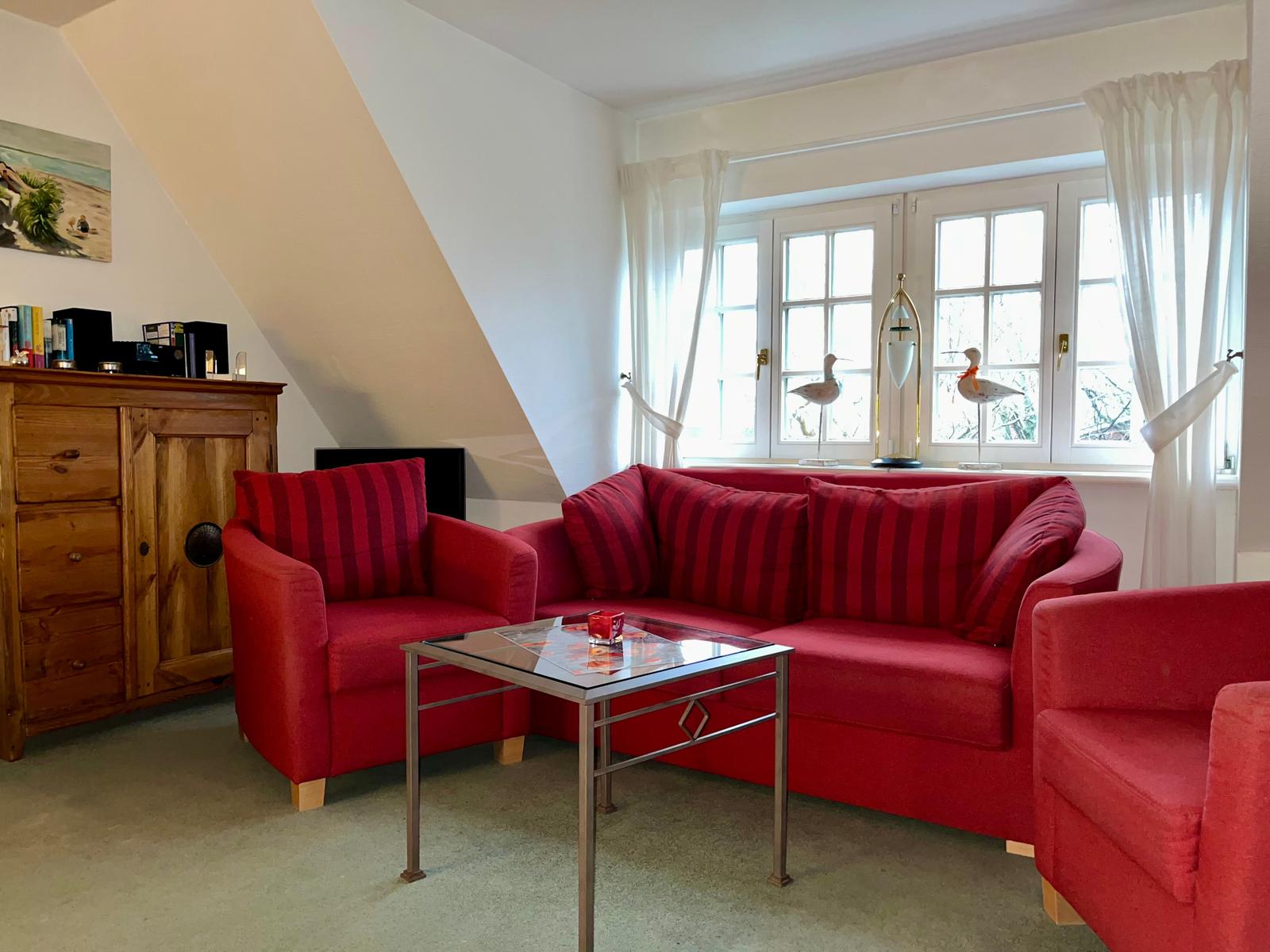 Wohnzimmer mit rotem Sofa, zwei Sesseln und Glas-Tisch. Fenster mit weißen Vorhängen.