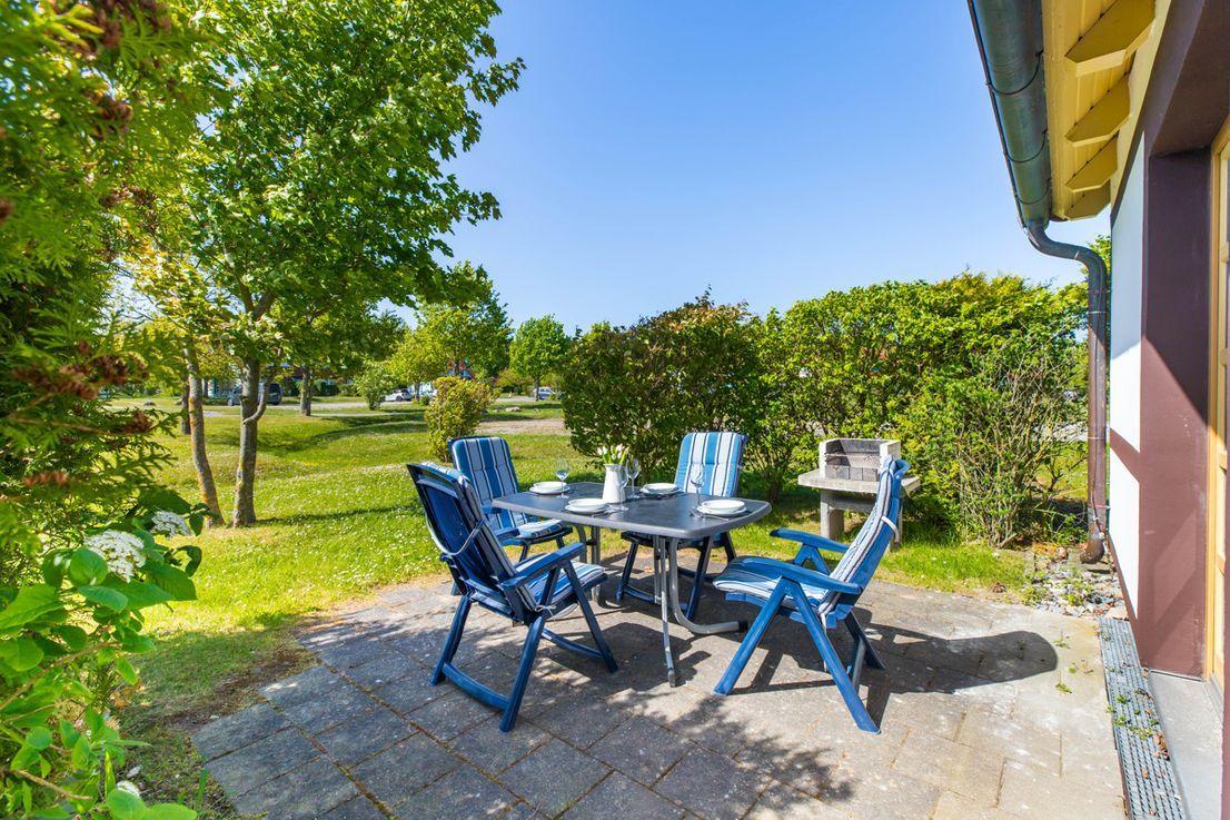 Terrasse mit Tisch, vier Stühlen und Grill. Grünfläche und Bäume im Hintergrund.