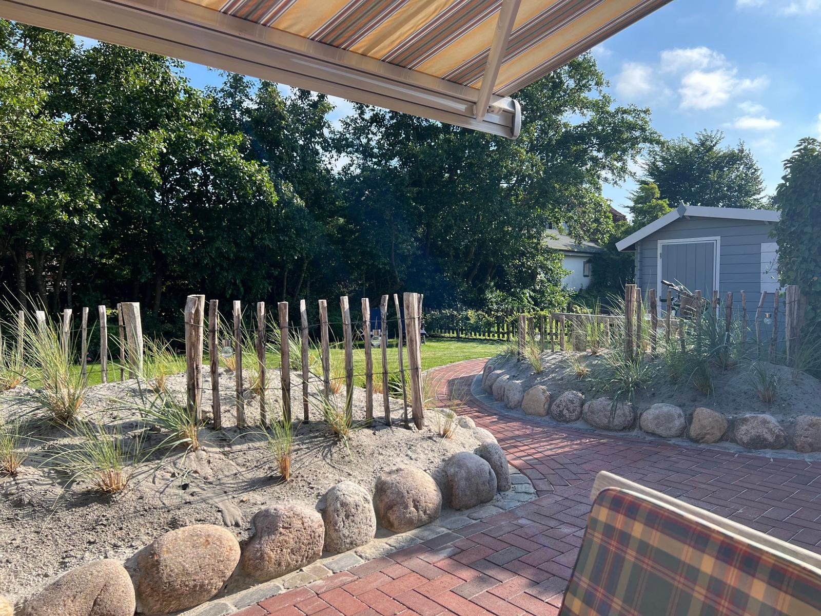 Terrasse mit Sonnenschutz, Sandbeete mit Gräsern und Steinumrandung, Pflasterweg und kleiner Gartenhaus im Hintergrund.