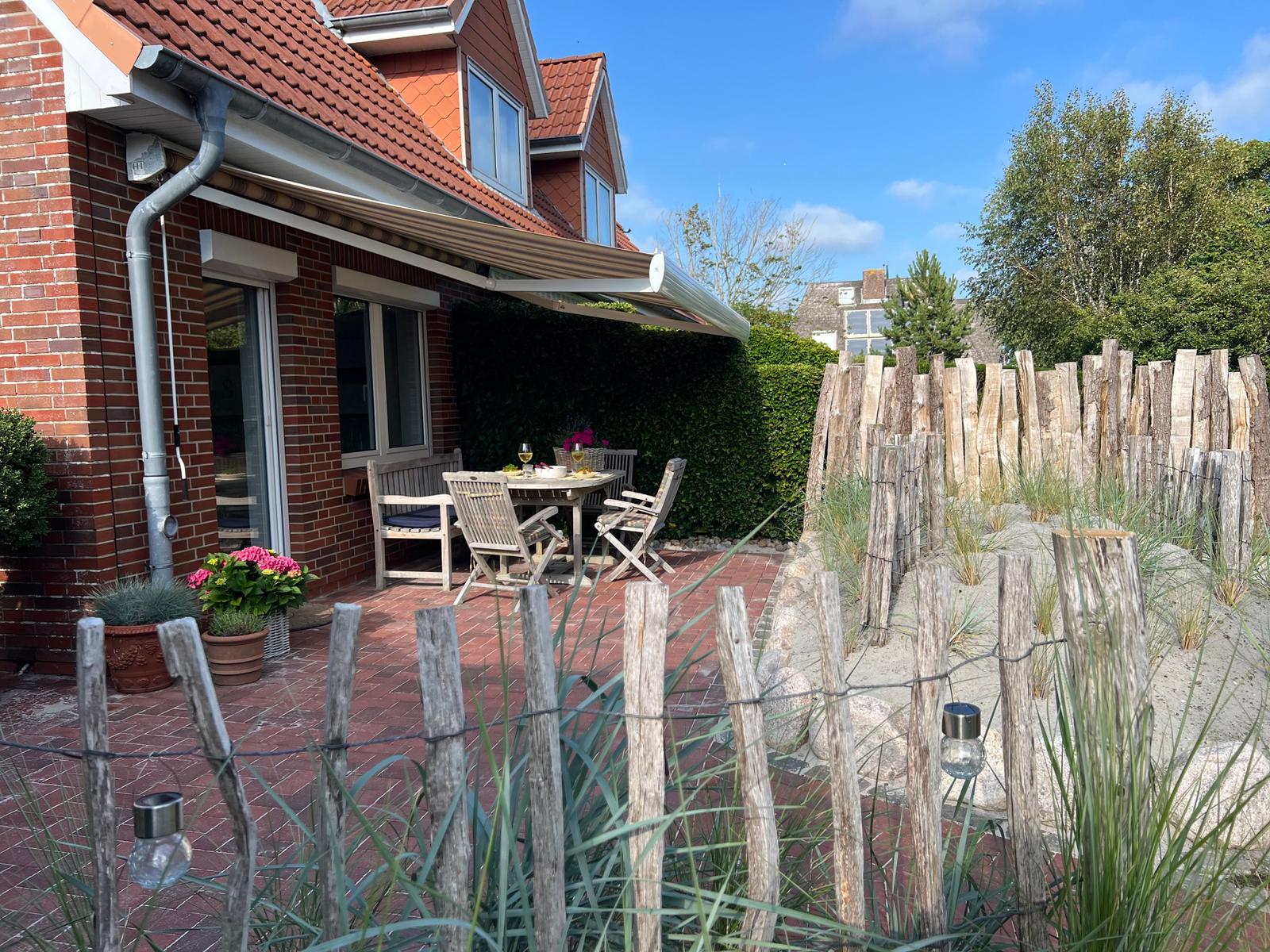 Terrasse mit Tisch, Stühlen und Markisen. Holzzaun und Sandstrand im Hintergrund.