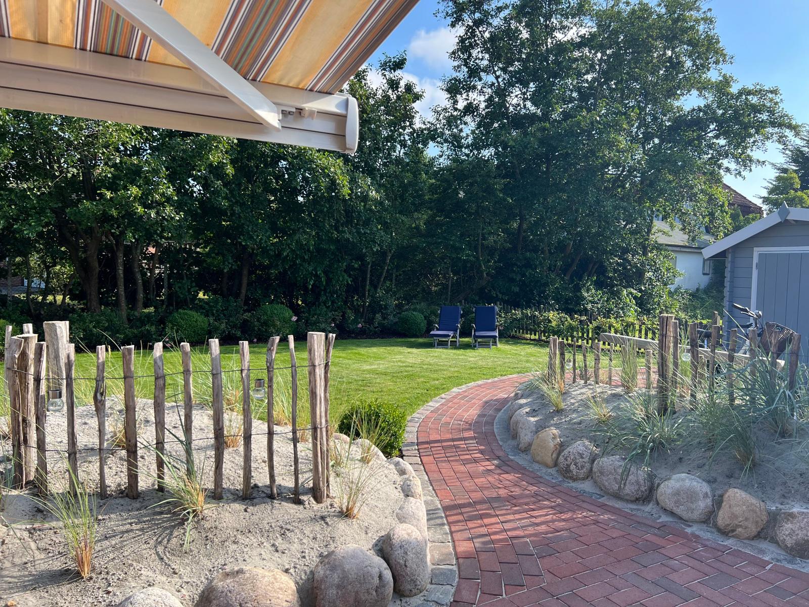Garten mit Steinterrasse, Holzzaun und Liegestühlen unter Bäumen.