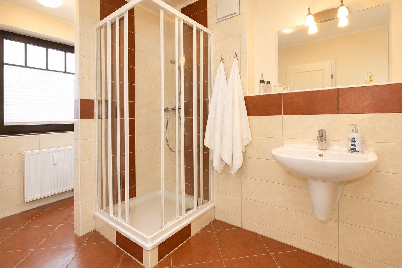 Badezimmer mit Dusche, Waschbecken und Fenster. Weiße Handtücher hängen an der Wand.