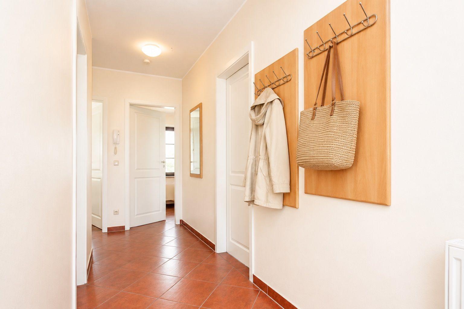 Flur mit Holzgarderoben, Mantel und Tasche hängen, Terrakottafliesen und weiße Türen.