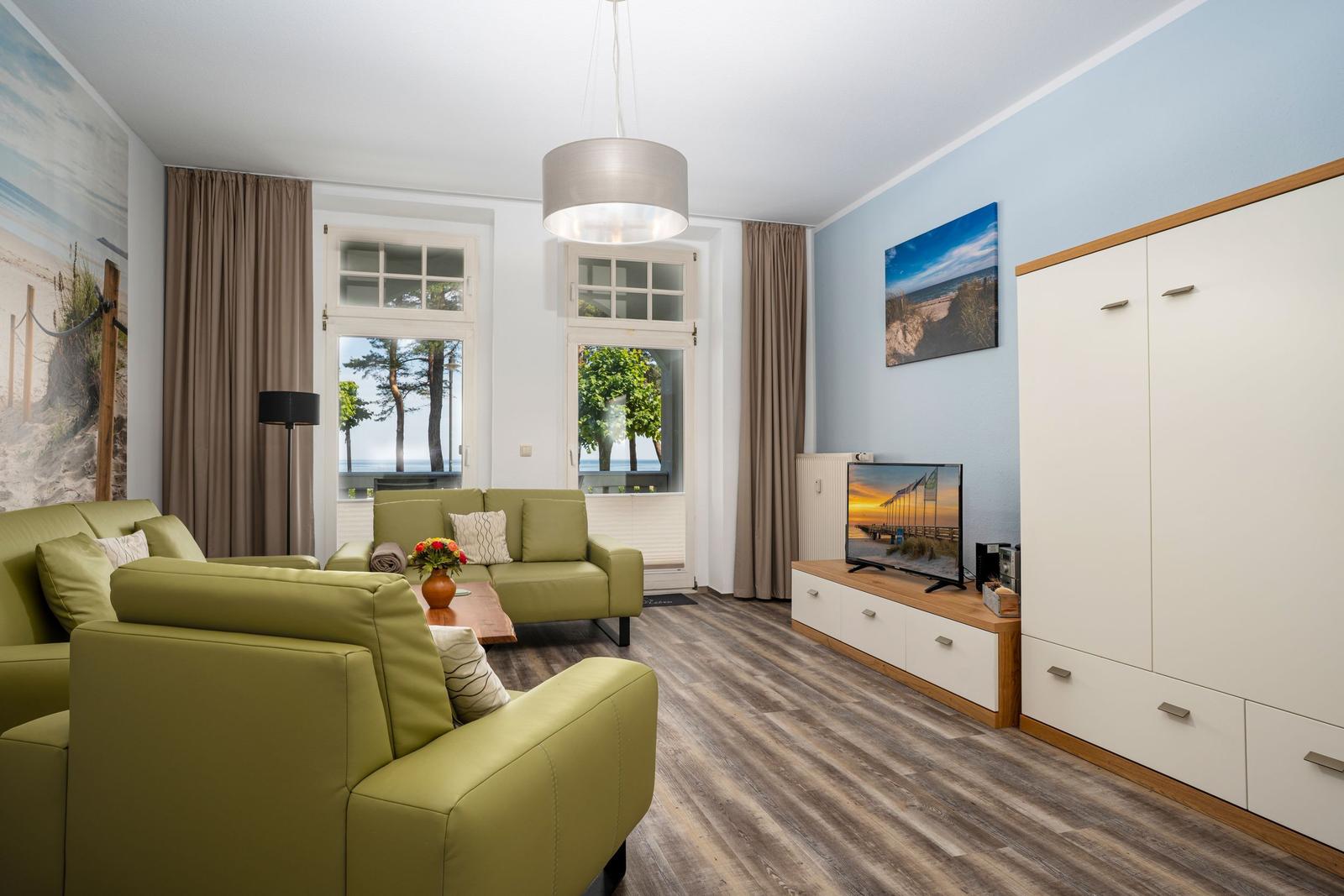Living room with green sofas, wood floor, and TV. Windows with view of trees.