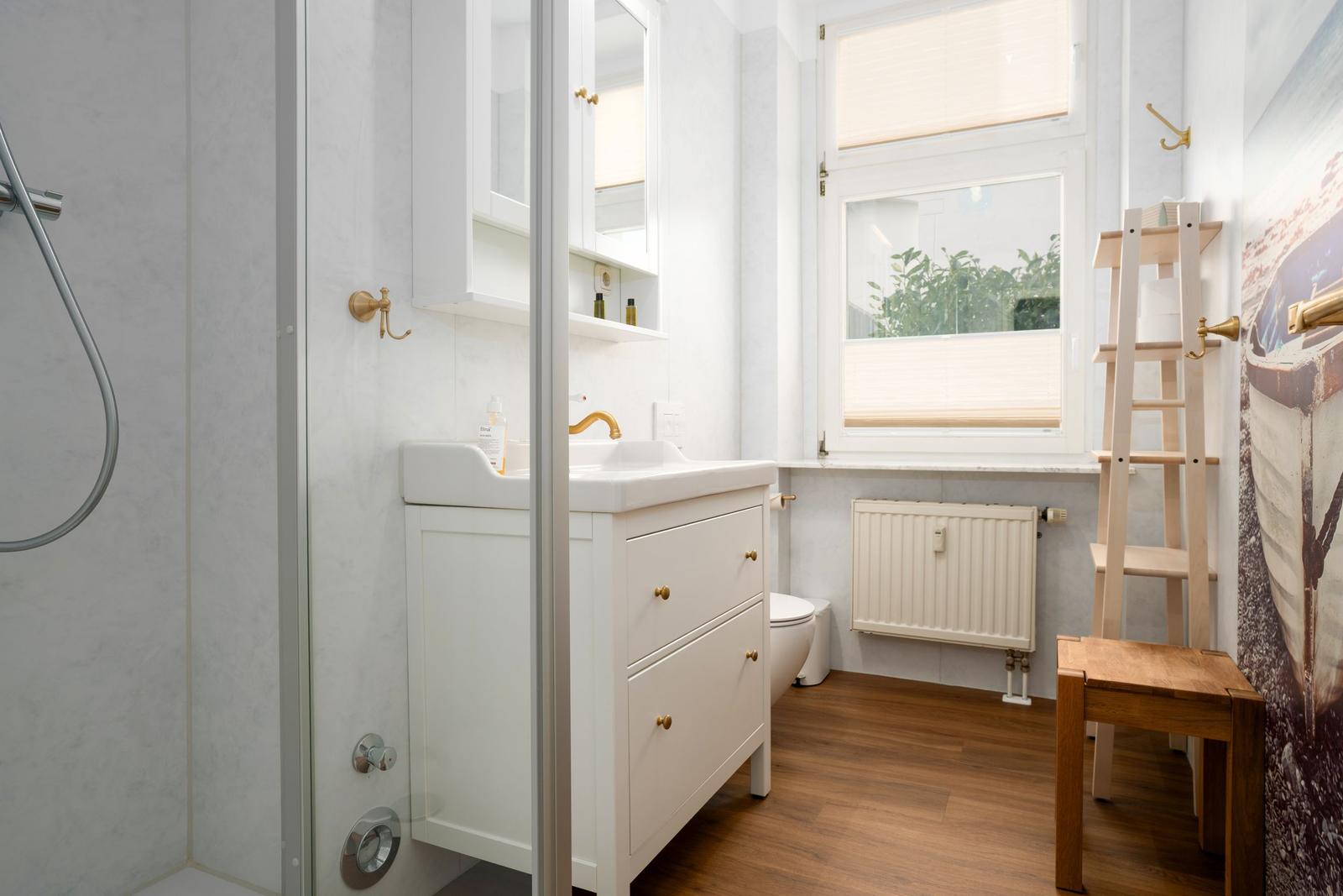 Bathroom with shower, sink, and toilet. Wooden floor and window with blinds.