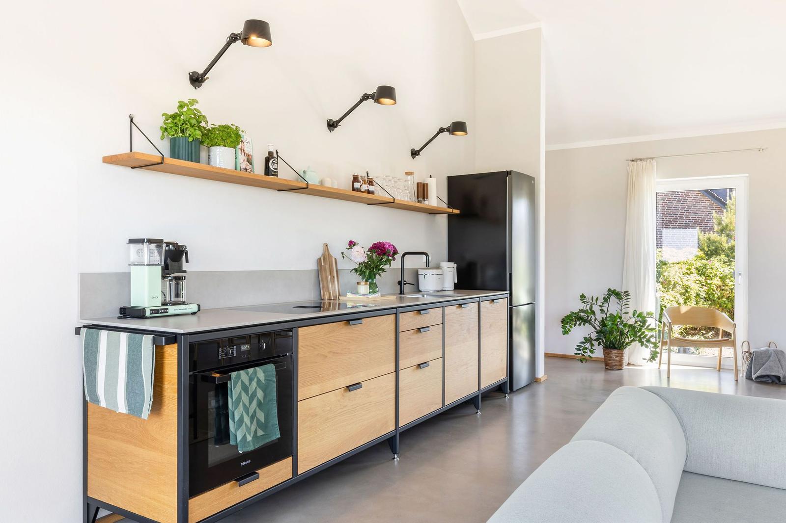 Moderne Küche mit Holzschränken, Kühlschrank und Arbeitsfläche. Wohnbereich mit Sofa und Pflanzen.