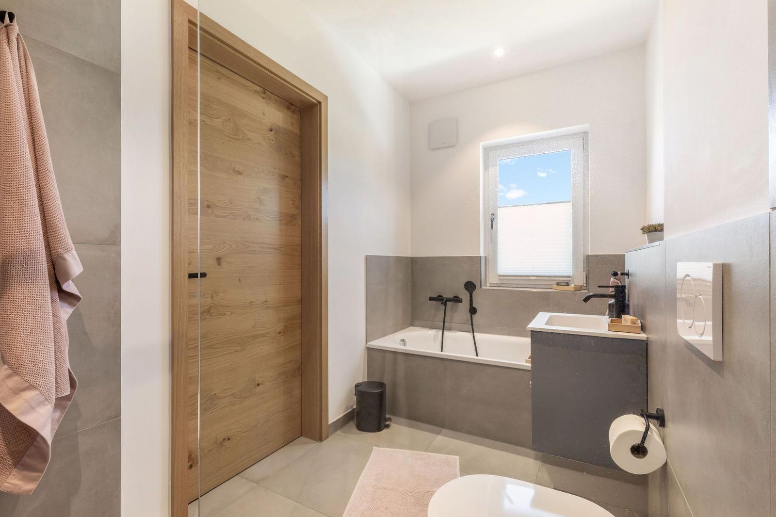 Bad mit Badewanne, Waschbecken und Toilette. Holztür und Fenster mit Jalousie.