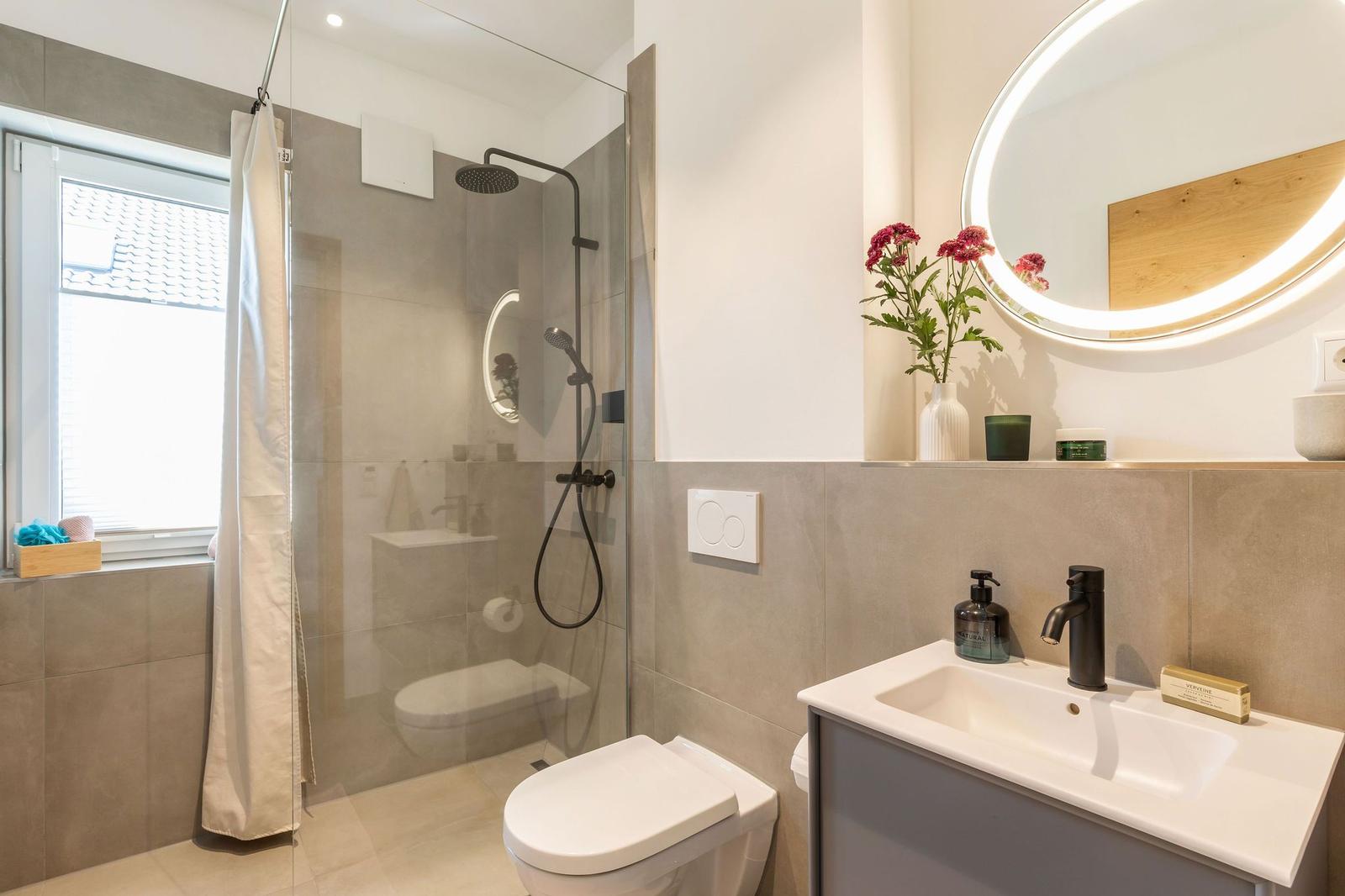Badezimmer mit Dusche, Waschbecken und Toilette. Rundes Spiegel mit Beleuchtung und Blumen.