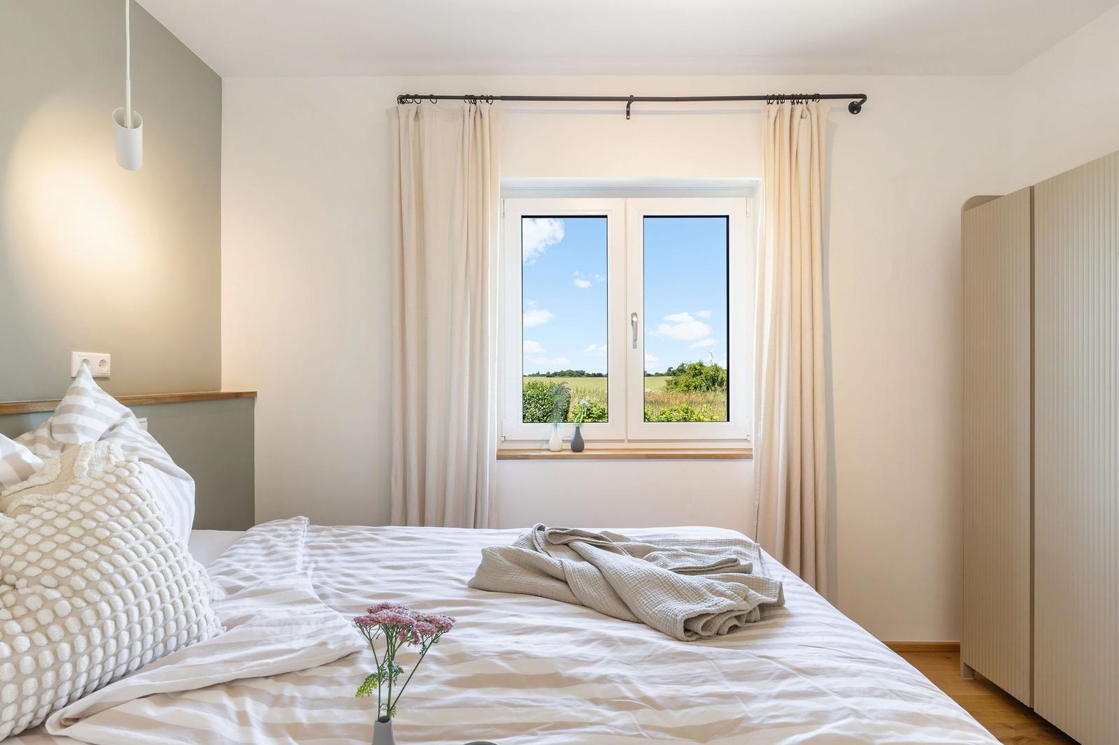 Schlafzimmer mit Bett, Fenster und Kleiderschrank. Draußen ist ein Feld mit blauem Himmel.