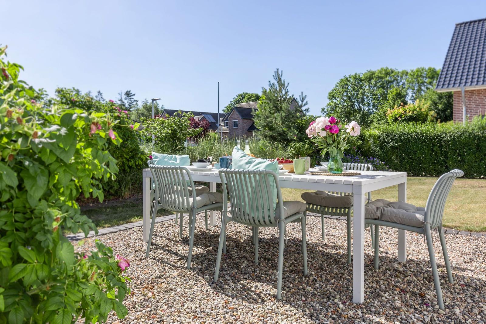 Ein weißer Tisch mit Stühlen steht auf Kies im Garten. Blumen und Obst sind darauf platziert.