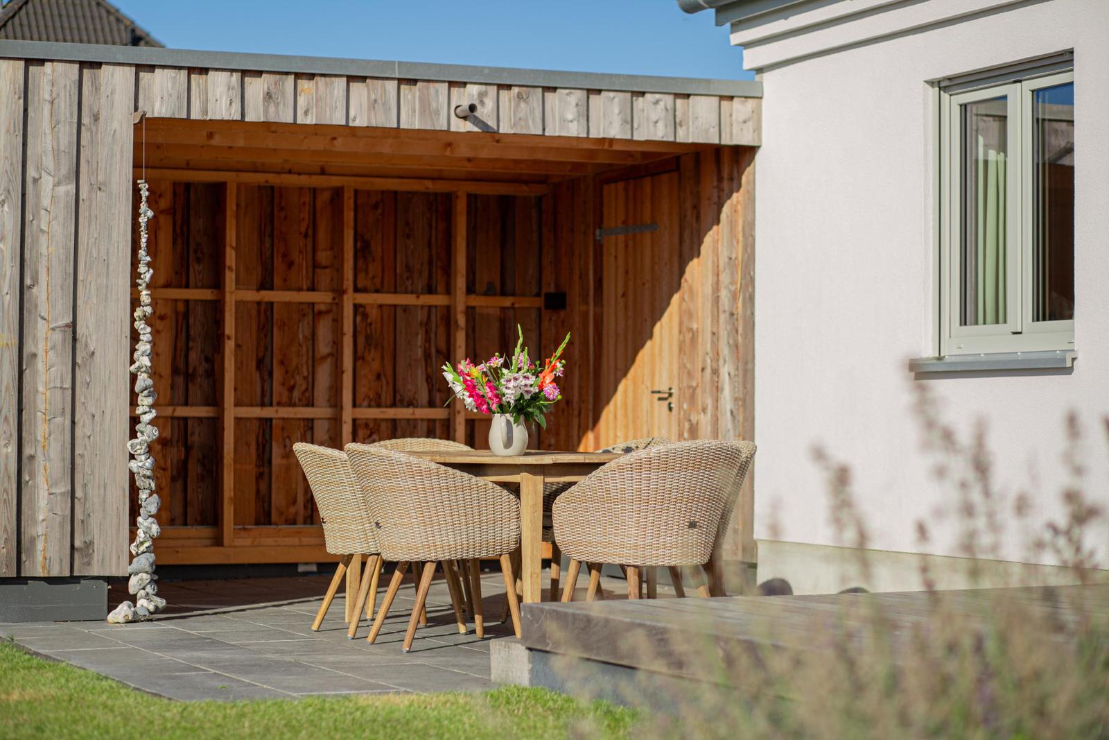 Holzterrasse mit Tisch, Stühlen und Blumenarrangement vor einem Gartenhaus.