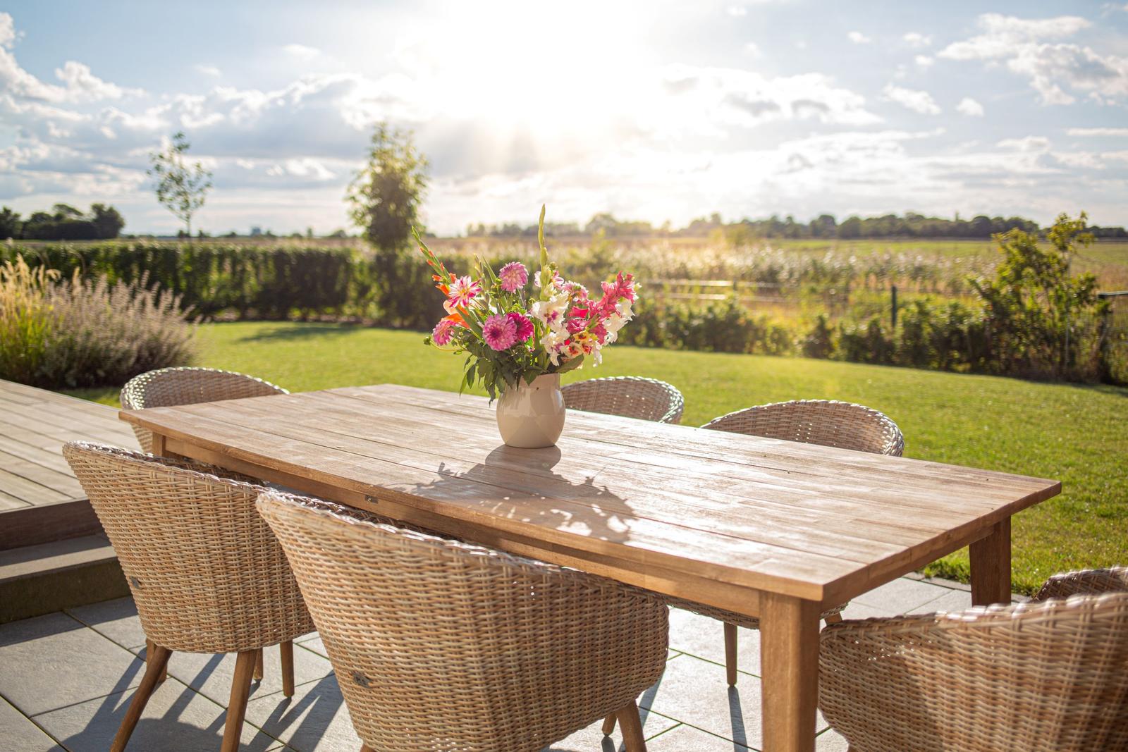 Holztisch mit Stühlen auf Terrasse, Blumenarrangement, Blick auf grünes Feld.