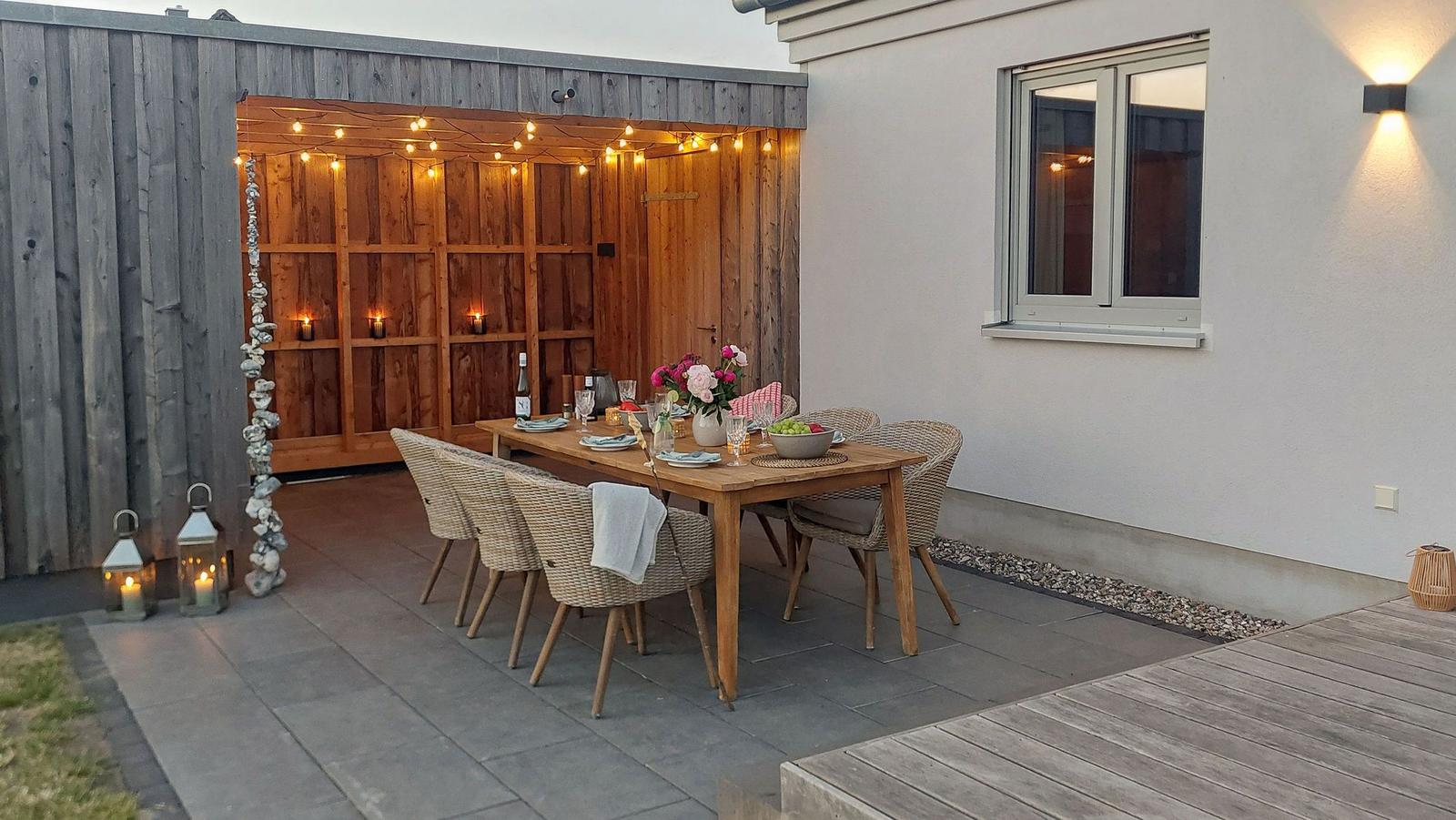 Holztisch mit Stühlen auf Terrasse, beleuchtet durch Girlanden und Laterne.