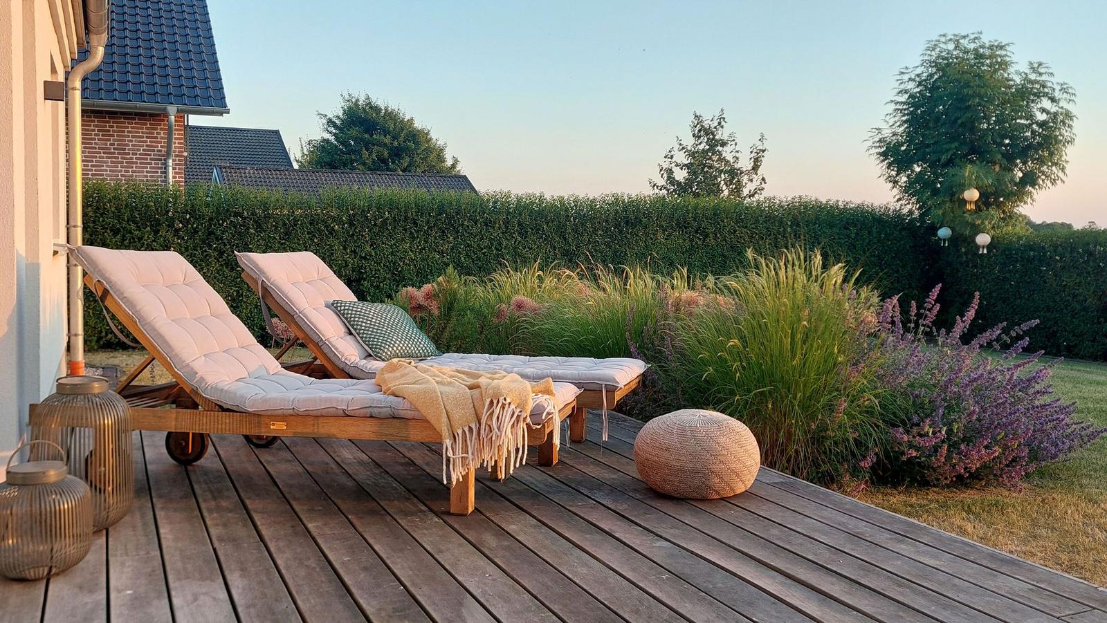 Terrasse mit zwei Liegestühlen, Decke und Kissen. Hintergrund: grüner Heckenzaun und Blumenbeete.