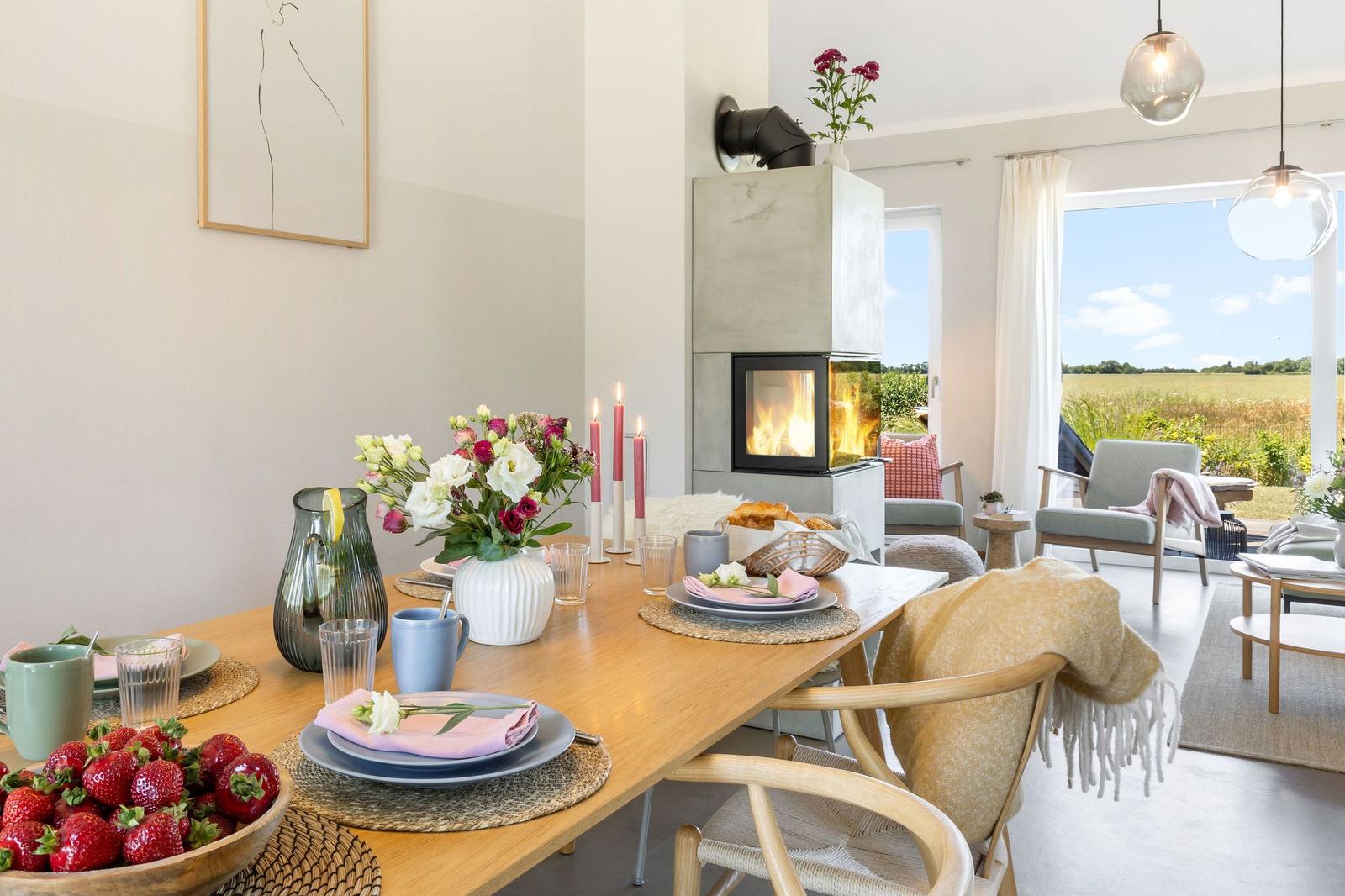 Esszimmer mit Holztisch, Blumen und Kamin. Blick auf Felder durch Fenster.
