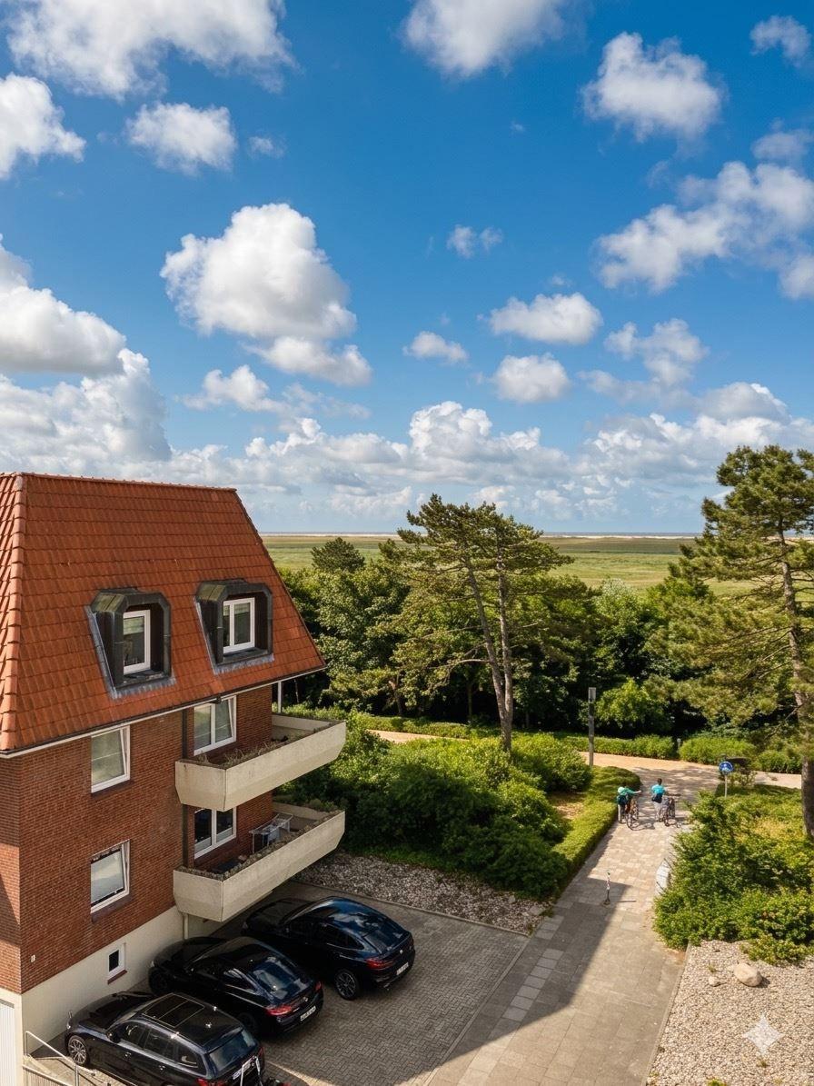 Haus mit rotem Dach, Balkonen und Parkplatz. Blick auf Wiesen und Bäume unter blauem Himmel mit weißen Wolken.