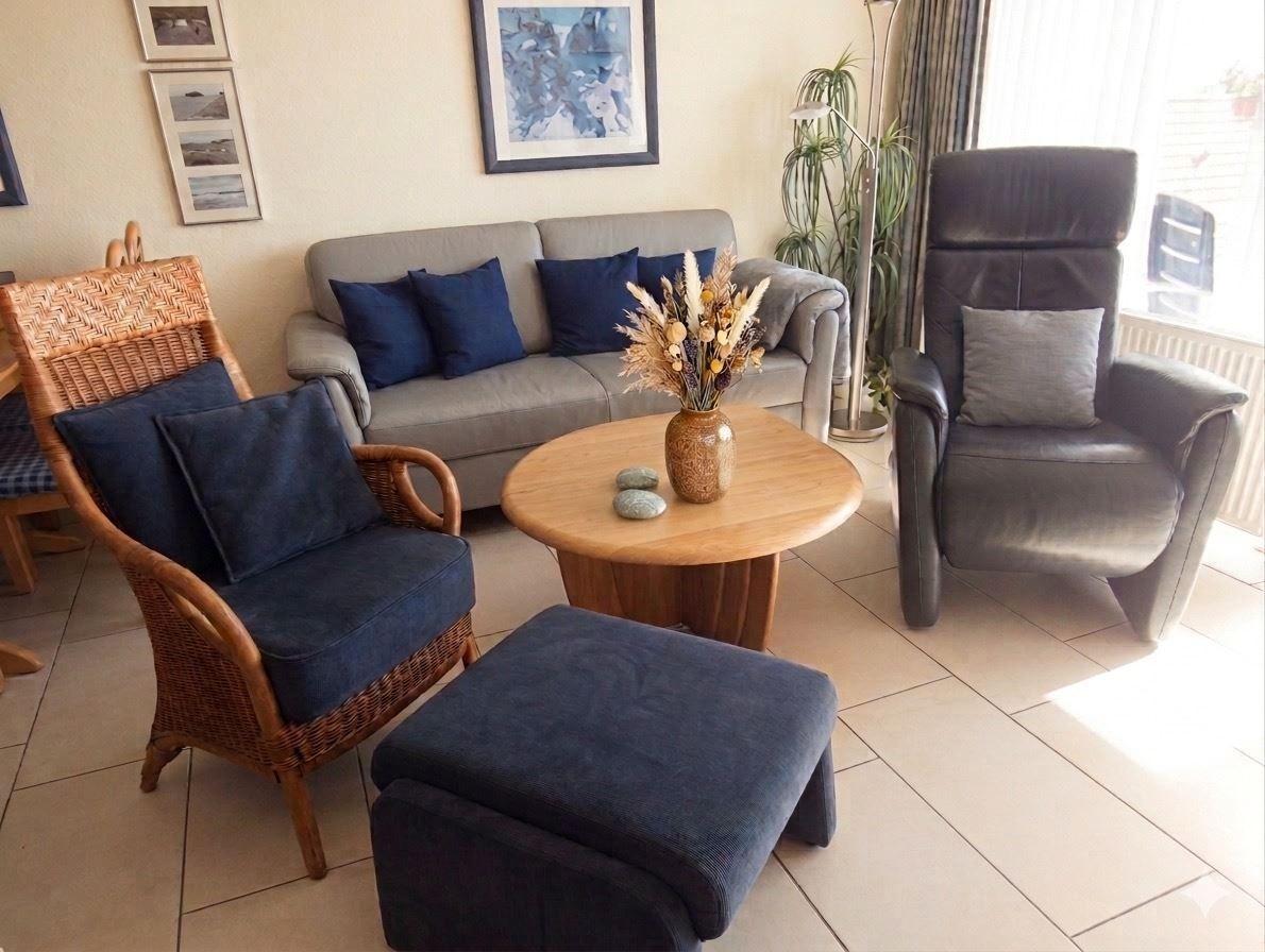 Wohnzimmer mit Sofa, Sessel und runden Tisch. Auf dem Tisch steht eine Vase mit Trockenblumen.