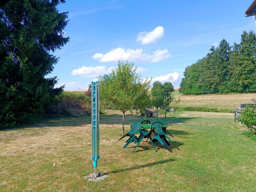 Green garden with table, chairs, and umbrella. Background: meadow and trees.