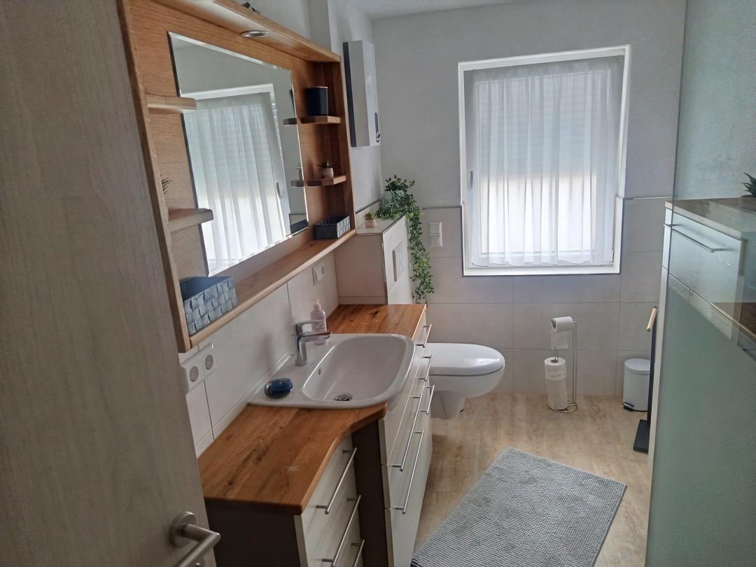 Bathroom with sink, toilet, and shower. Wooden countertop and window with curtains.