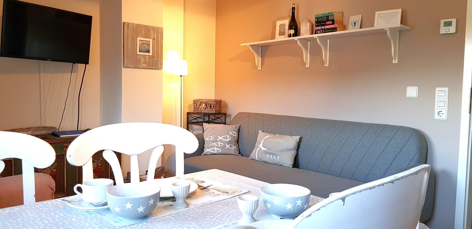 Grauer Sofa mit Kissen, Tisch mit Tassen und Schalen, Wandregal und Fernseher.