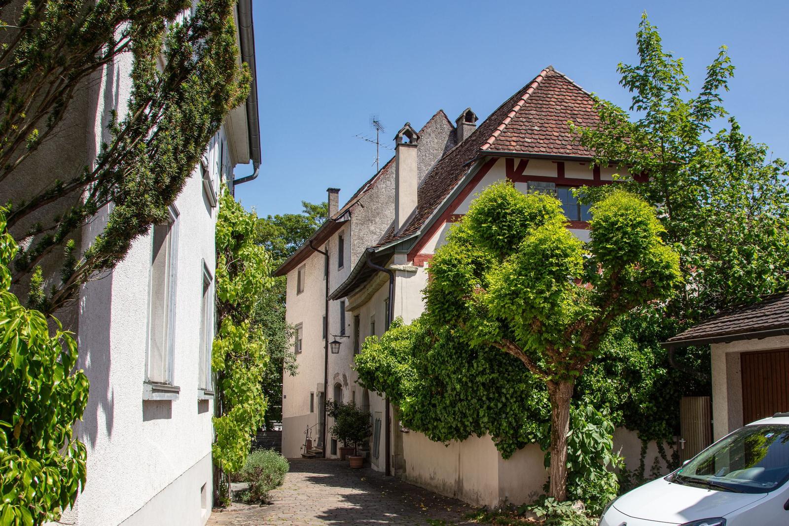 Enge Gasse mit traditionellen Häusern und grünen Bäumen unter blauem Himmel.