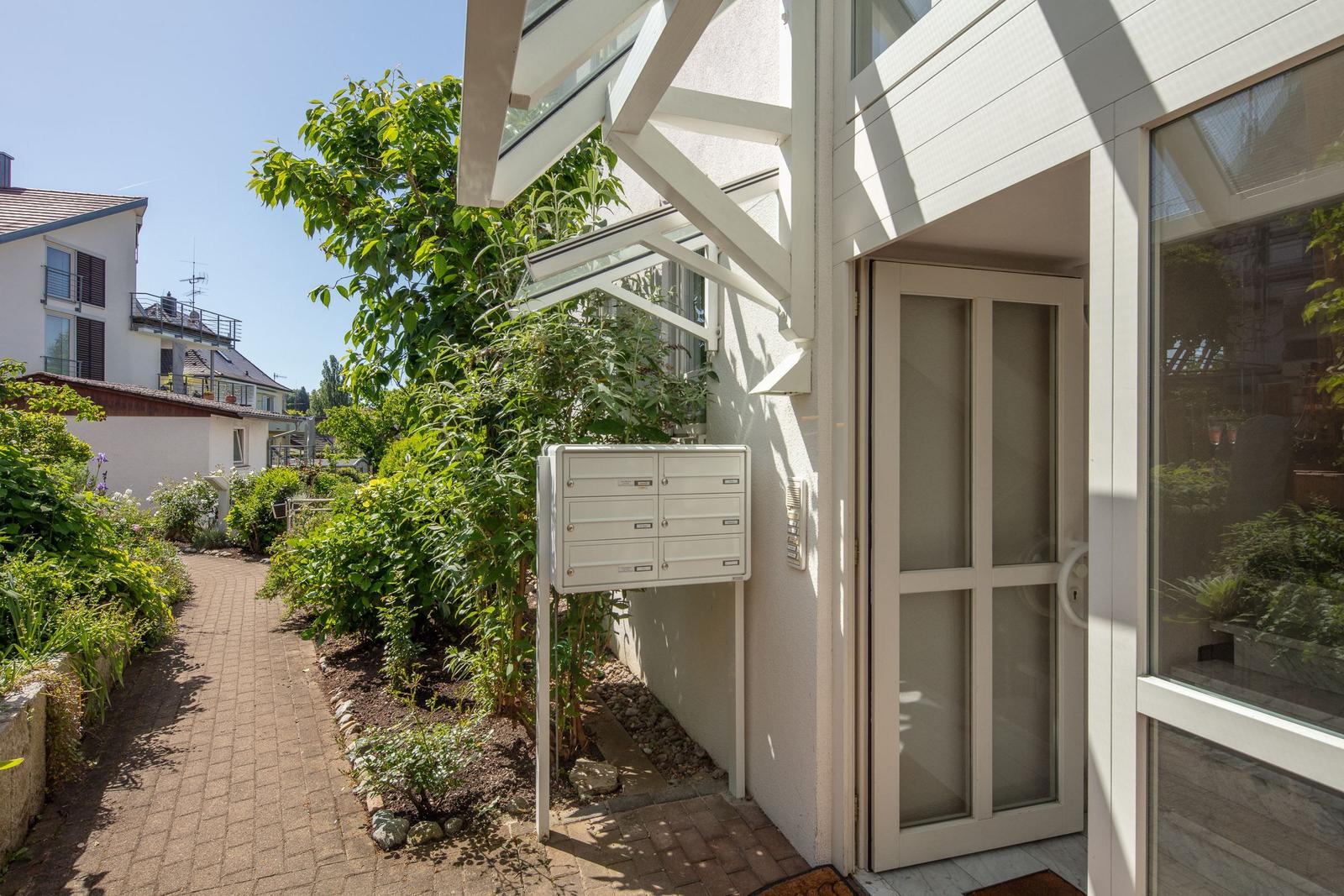 Eingangsbereich mit Glastür, Briefkasten und gepflasterter Weg durch grünen Garten.