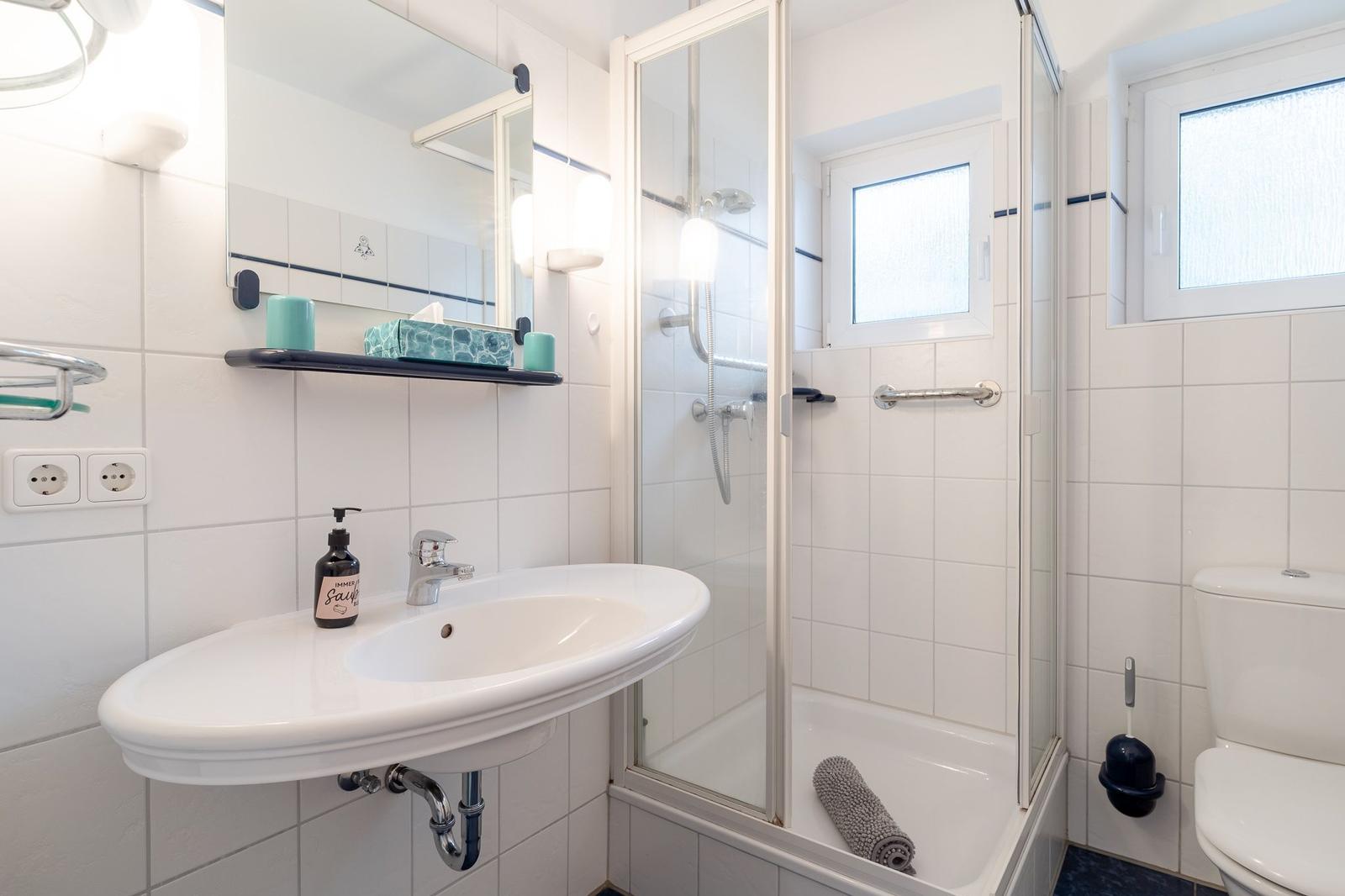 Badezimmer mit Dusche, Waschbecken und Toilette. Weiße Fliesen und Fenster.