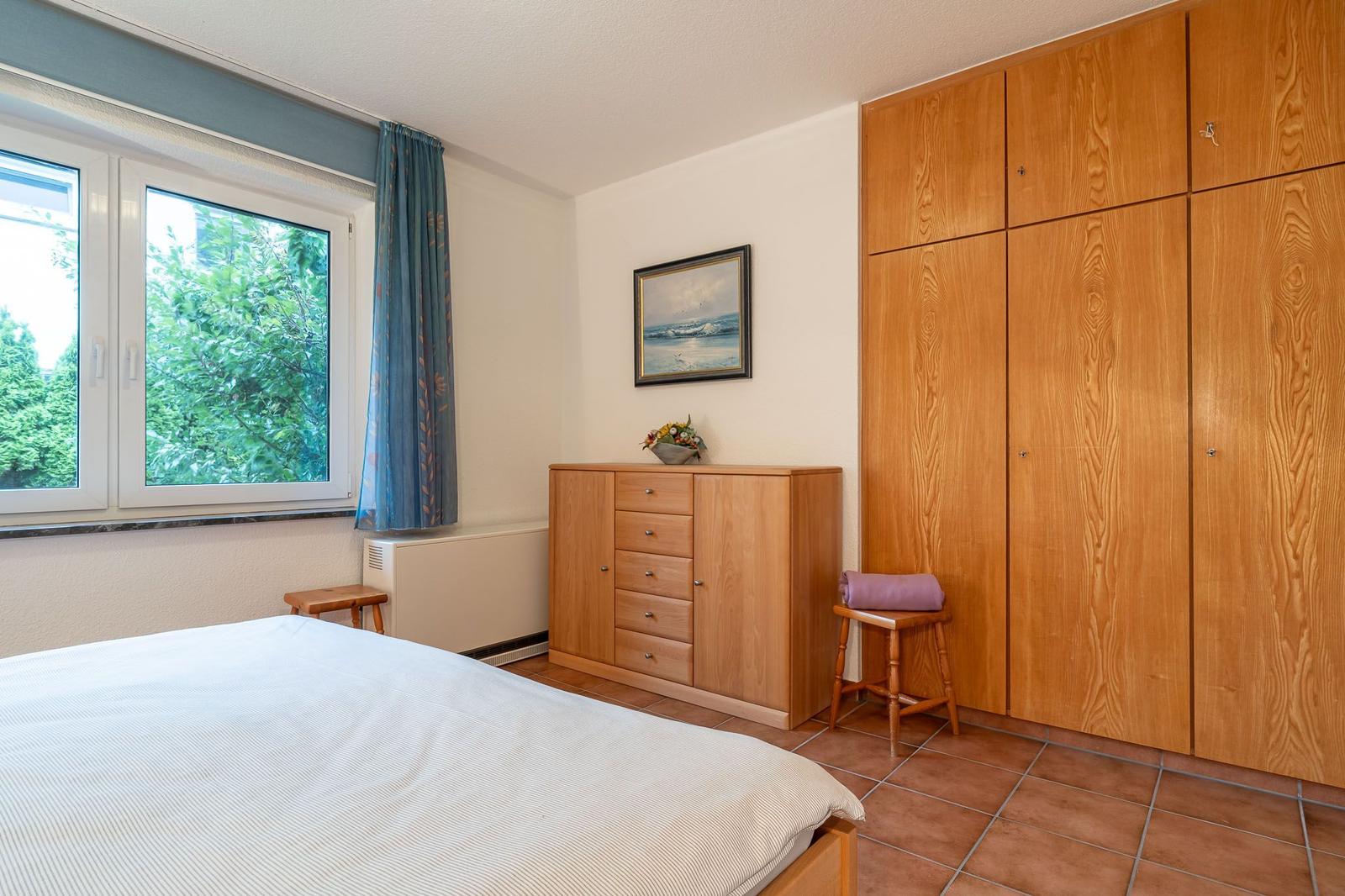 Schlafzimmer mit Bett, Kleiderschrank und Fenster mit Blick auf Bäume.
