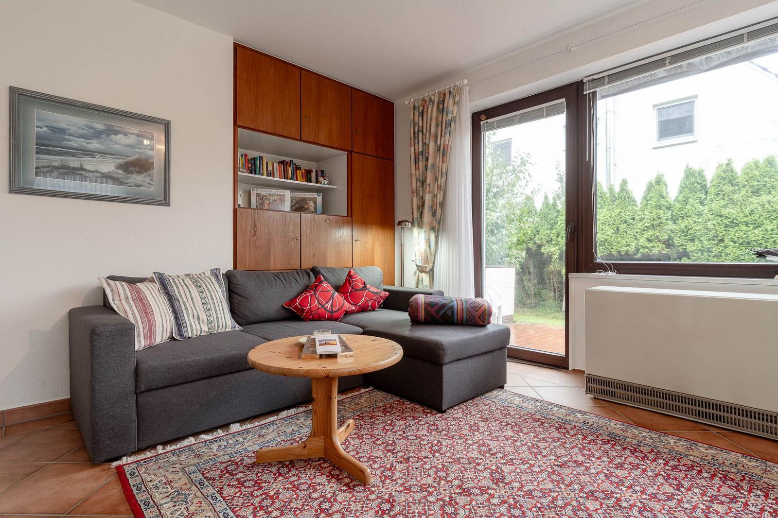 Wohnzimmer mit grauem Sofa, Holztisch und rotem Teppich. Große Fenster und Bücherregal.