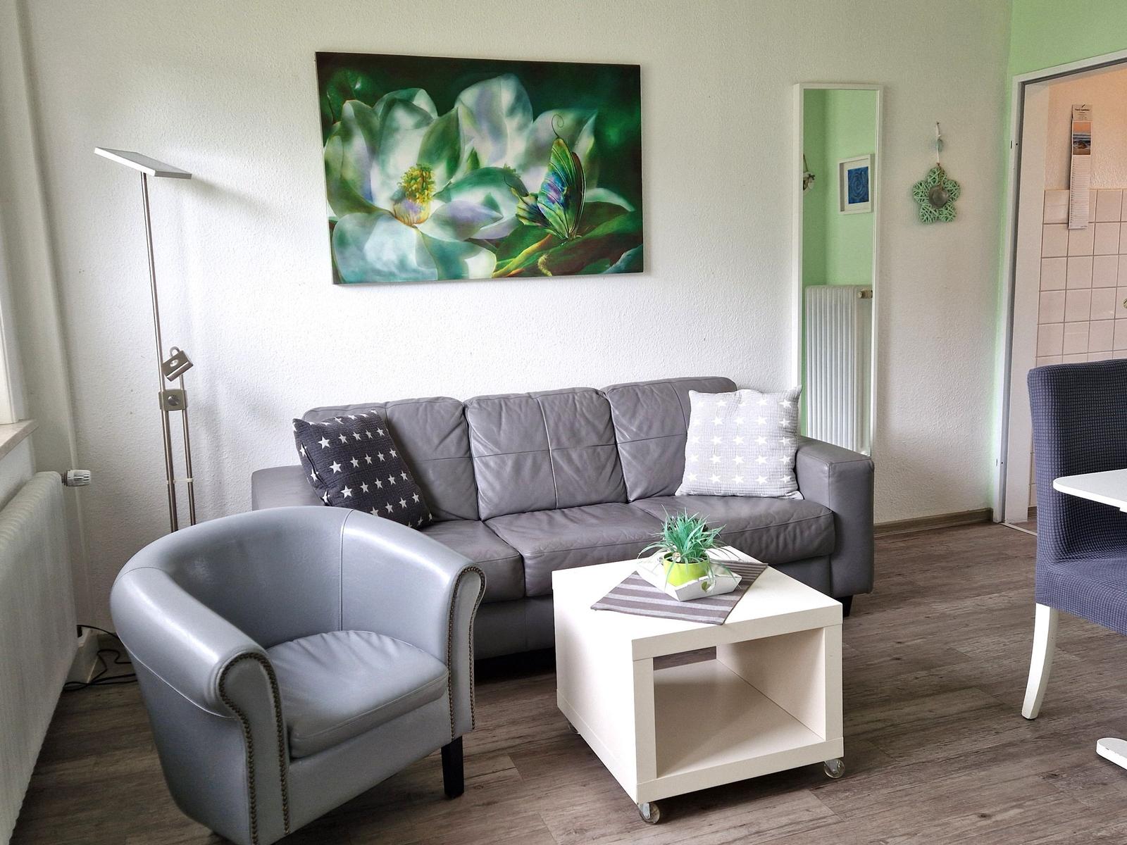 Ein Wohnzimmer mit grauem Sofa, einem Sessel und einem Tisch. An der Wand hängt ein Bild.